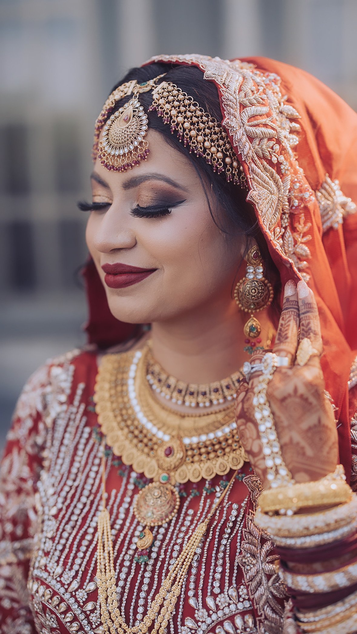 A woman in traditional bridal attire, featuring intricate jewelry, a henna design on her hand, and an embroidered red veil.