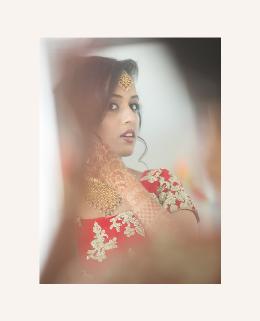 A woman in traditional Indian attire and jewelry, with henna on her hand, looking at her reflection.