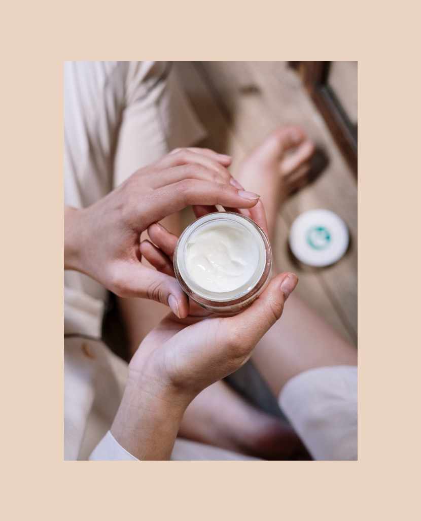 Person holding a jar of cream with lid removed, sitting cross-legged on wooden floor.