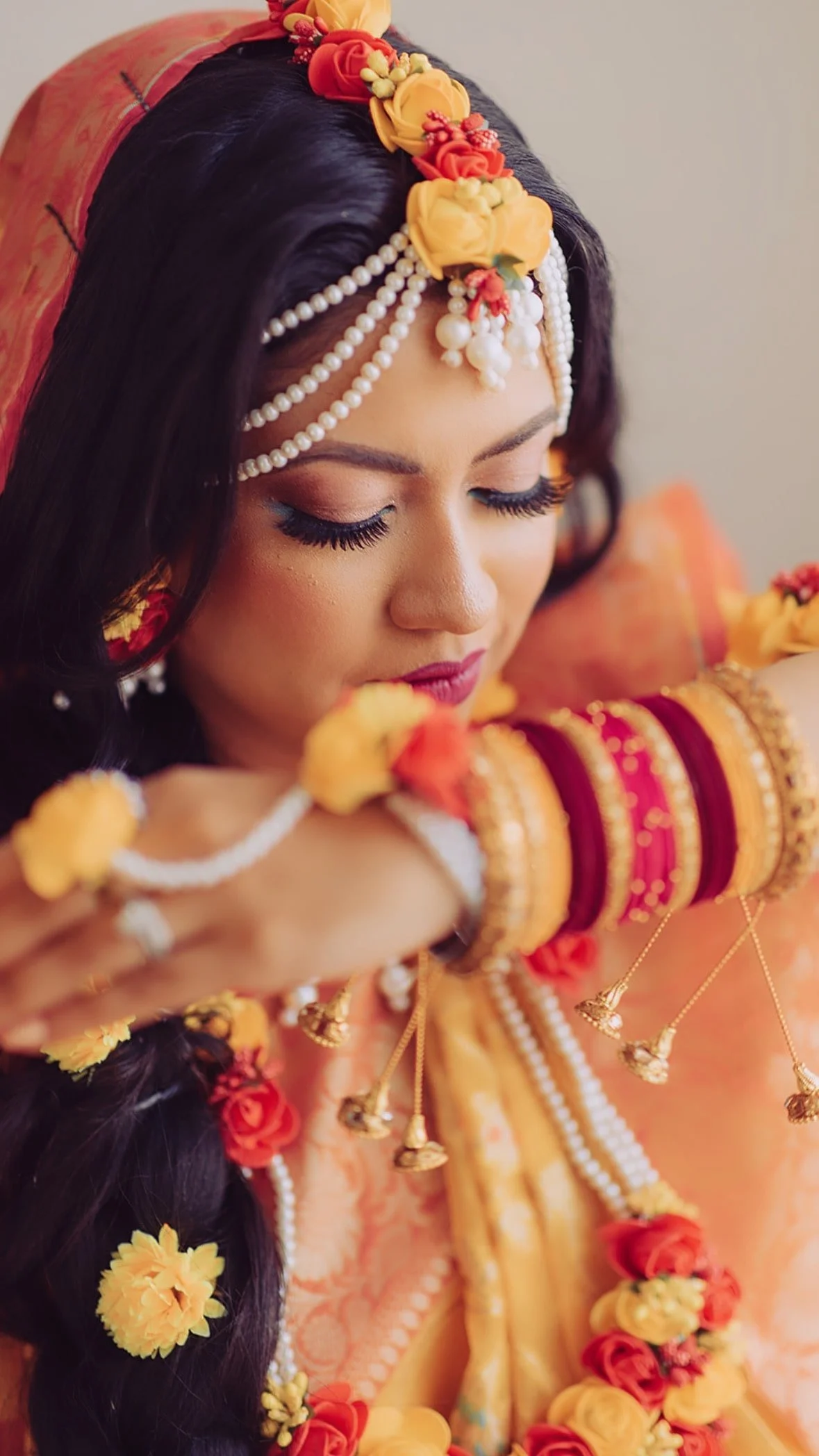 Woman in traditional Indian attire with floral and pearl jewelry, wearing red and yellow bangles, and adorned with flowers in her hair.