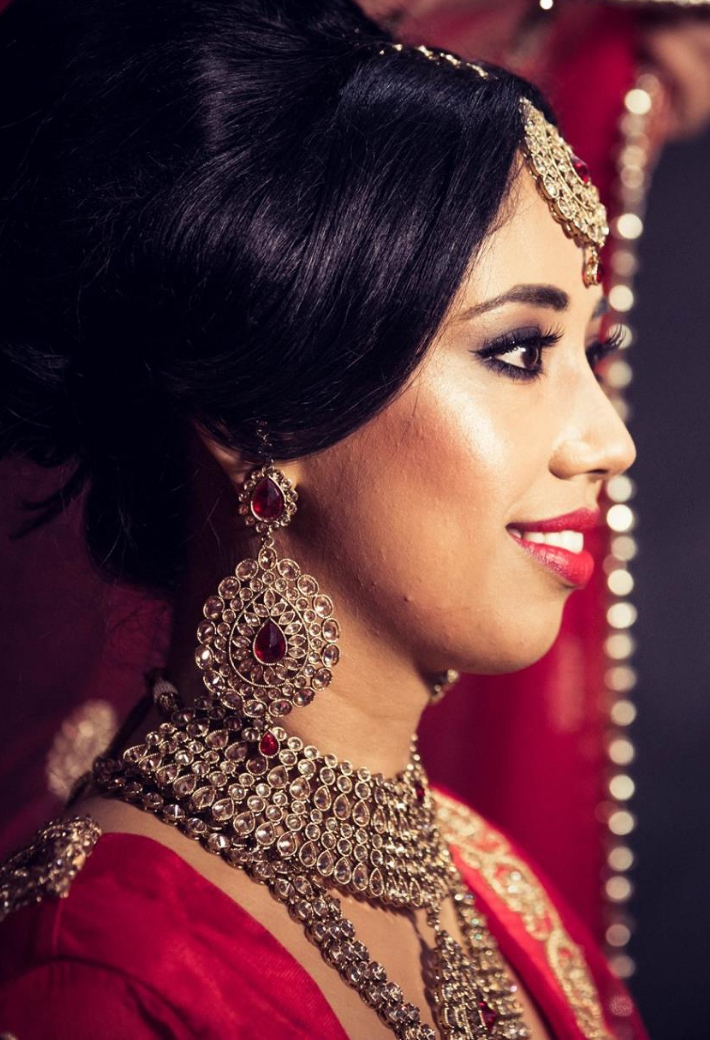 Close-up of a woman in traditional South Asian bridal attire, wearing an ornate necklace, earrings, and headpiece, with red and gold accents.