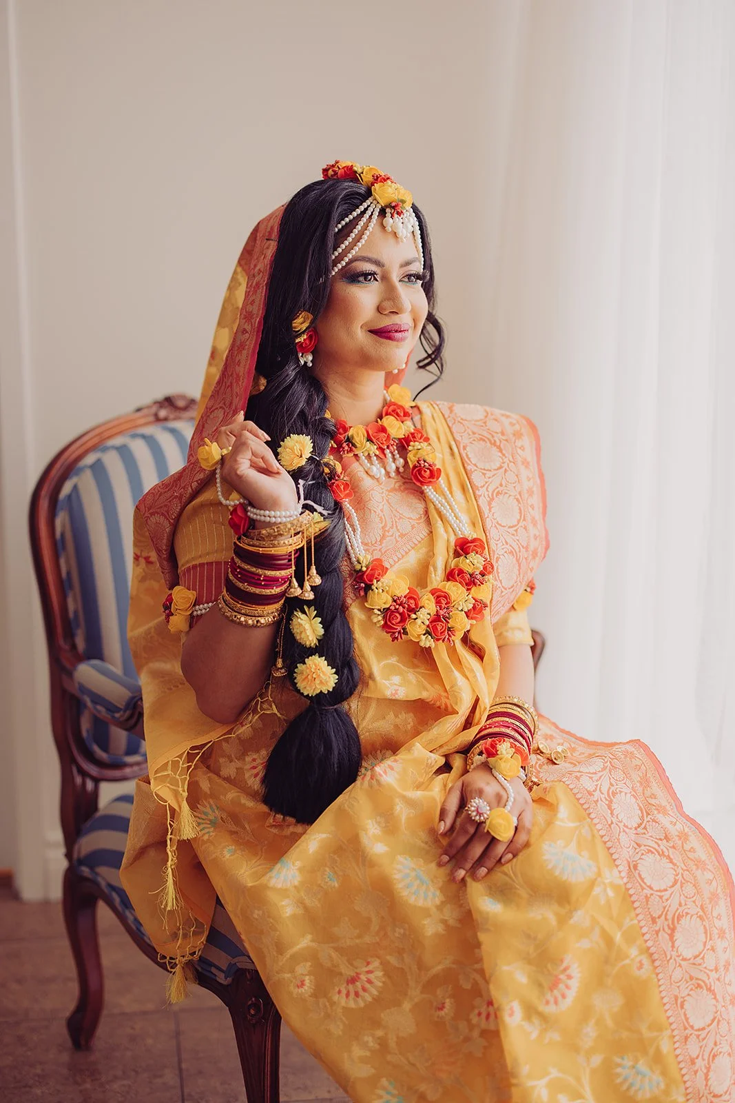 A woman dressed in traditional South Asian attire, wearing a yellow and orange sari with floral jewelry, sitting on a striped armchair. She has a floral headpiece, necklaces, and bangles, and appears to be preparing for a cultural ceremony or celebration.