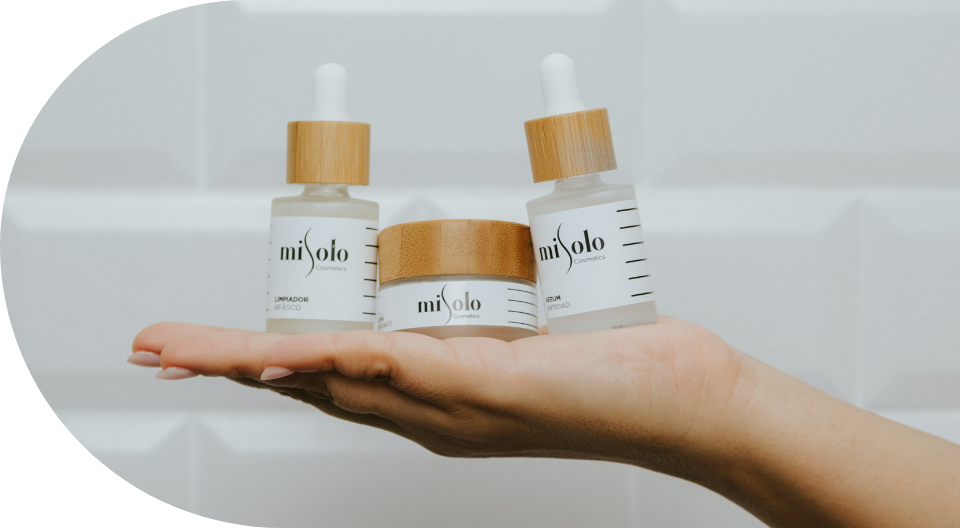 Hand holding three miSolo Cosmetics products with wooden caps, including two bottles and one jar, against a white tile background.
