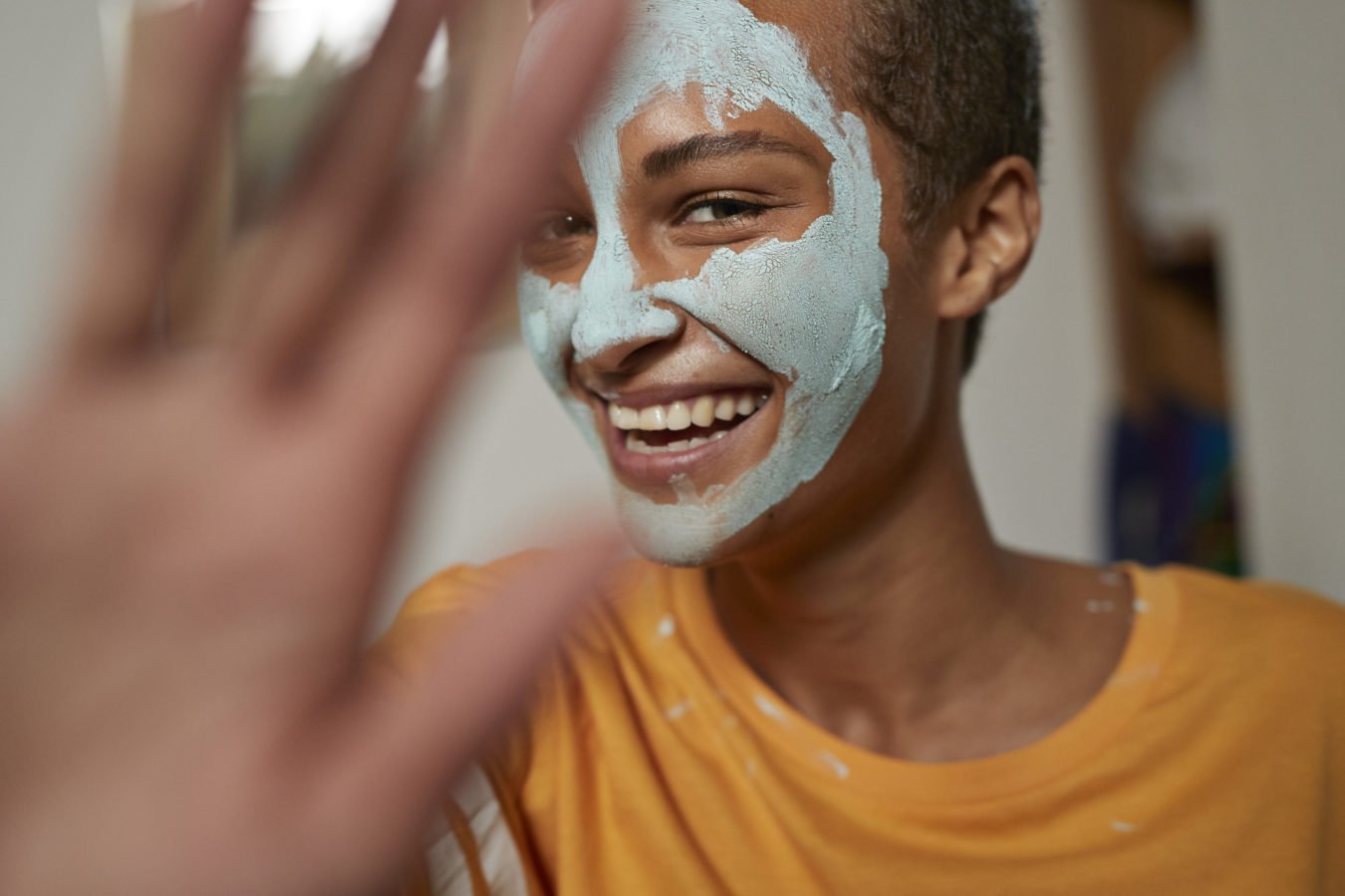 Person smiling with a facial mask on, wearing an orange shirt, partially obscured by an outstretched hand.
