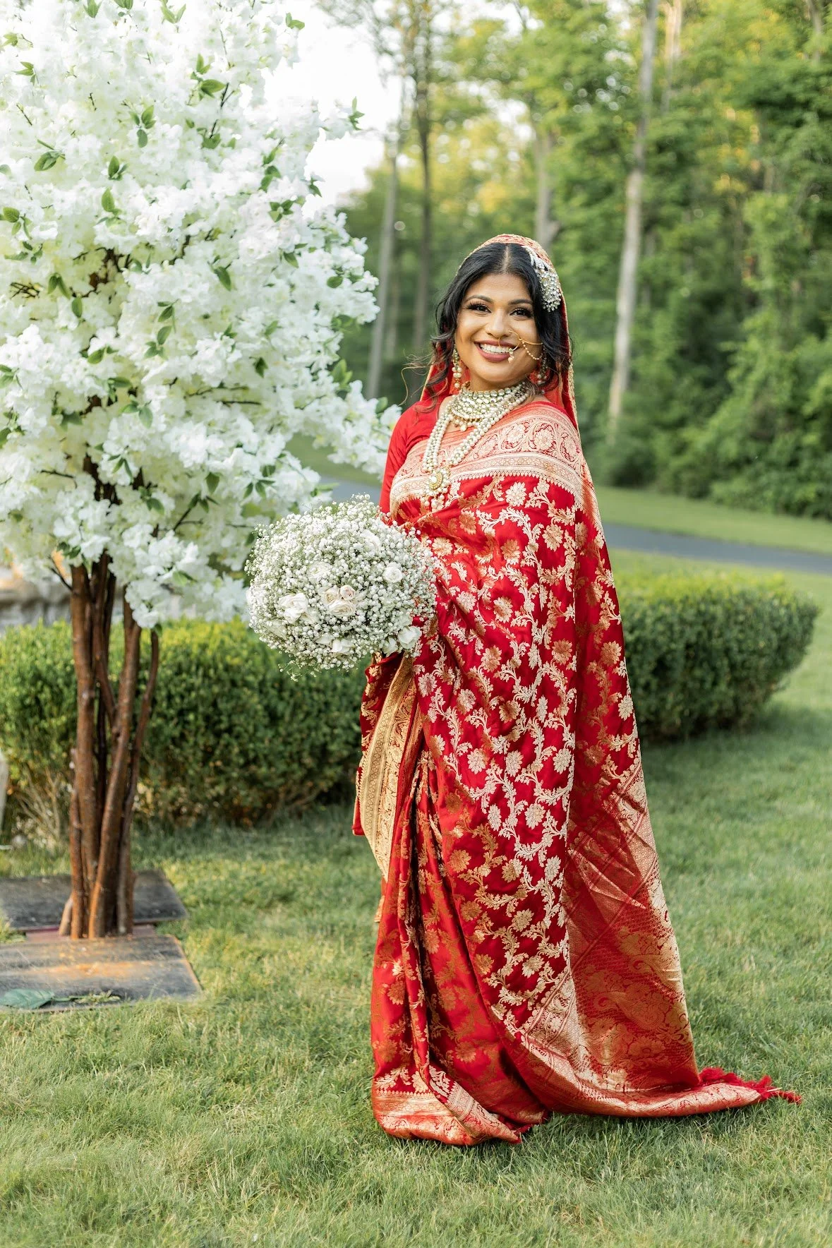 A woman in a red and gold traditional saree stands on grass beside a tree with white flowers, holding a bouquet of white flowers. She is smiling and has layered gold jewelry.
