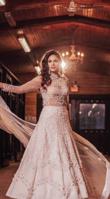 A woman in an ornate white wedding dress with long sleeves stands in a warmly lit hallway with a wooden ceiling. A chandelier is visible in the background.