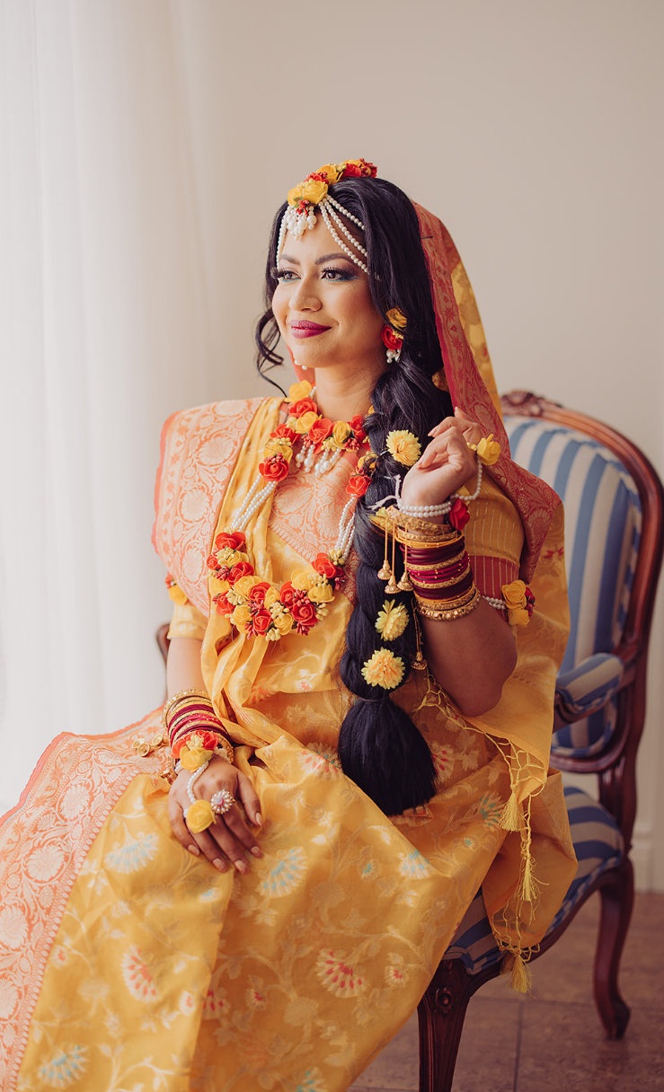A woman in traditional Indian attire sits on a striped chair, wearing a yellow and orange sari with floral jewelry and bangles. Her hair is adorned with flowers and she has a smile on her face.