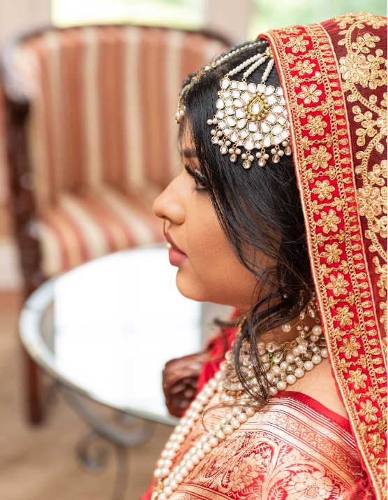 Side profile of a woman wearing traditional South Asian bridal attire, including a red embroidered veil and ornate jewelry, seated indoors.