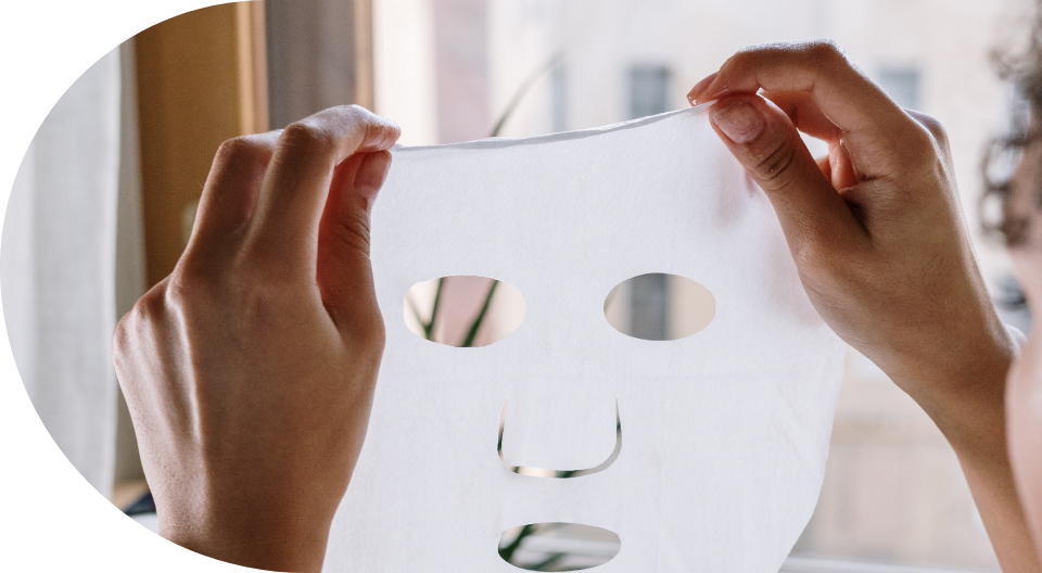 Person holding a white sheet face mask with cutouts for eyes, nose, and mouth near a window.