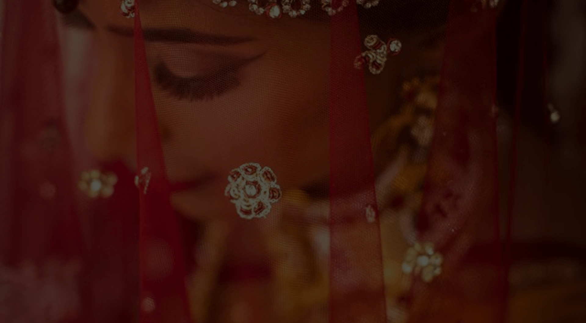 Close-up of a bride with red veil adorned with gold embroidery. Her eyes are closed with heavy eye makeup visible.