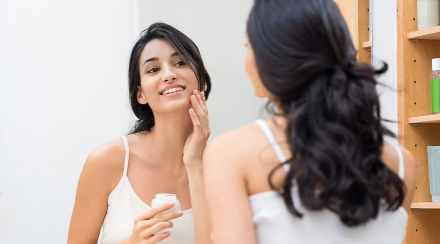 Woman applying moisturizer on face in front of bathroom mirror.