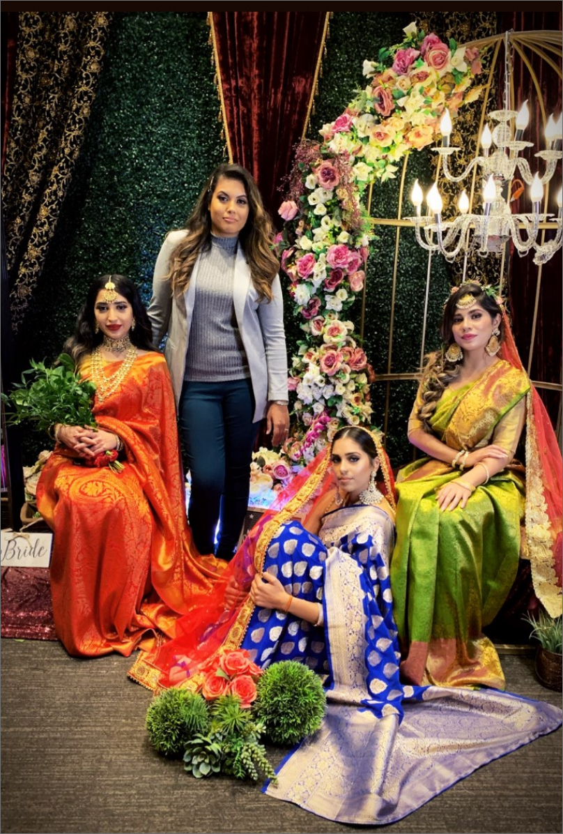 A group of four women posing indoors in front of a decorative floral backdrop. Three women are seated, wearing colorful traditional sarees in orange, blue, and green, with jewelry and headpieces. A fourth woman in a casual blazer and pants stands behind them. The setting is adorned with flowers and a lit chandelier.