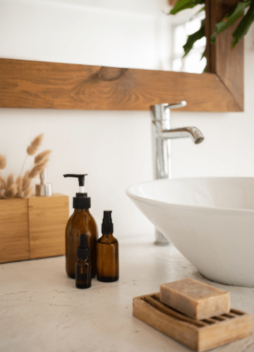 Bathroom countertop with a white sink, wooden mirror frame, amber glass bottles, soap dish with bar soap, and decorative reeds.