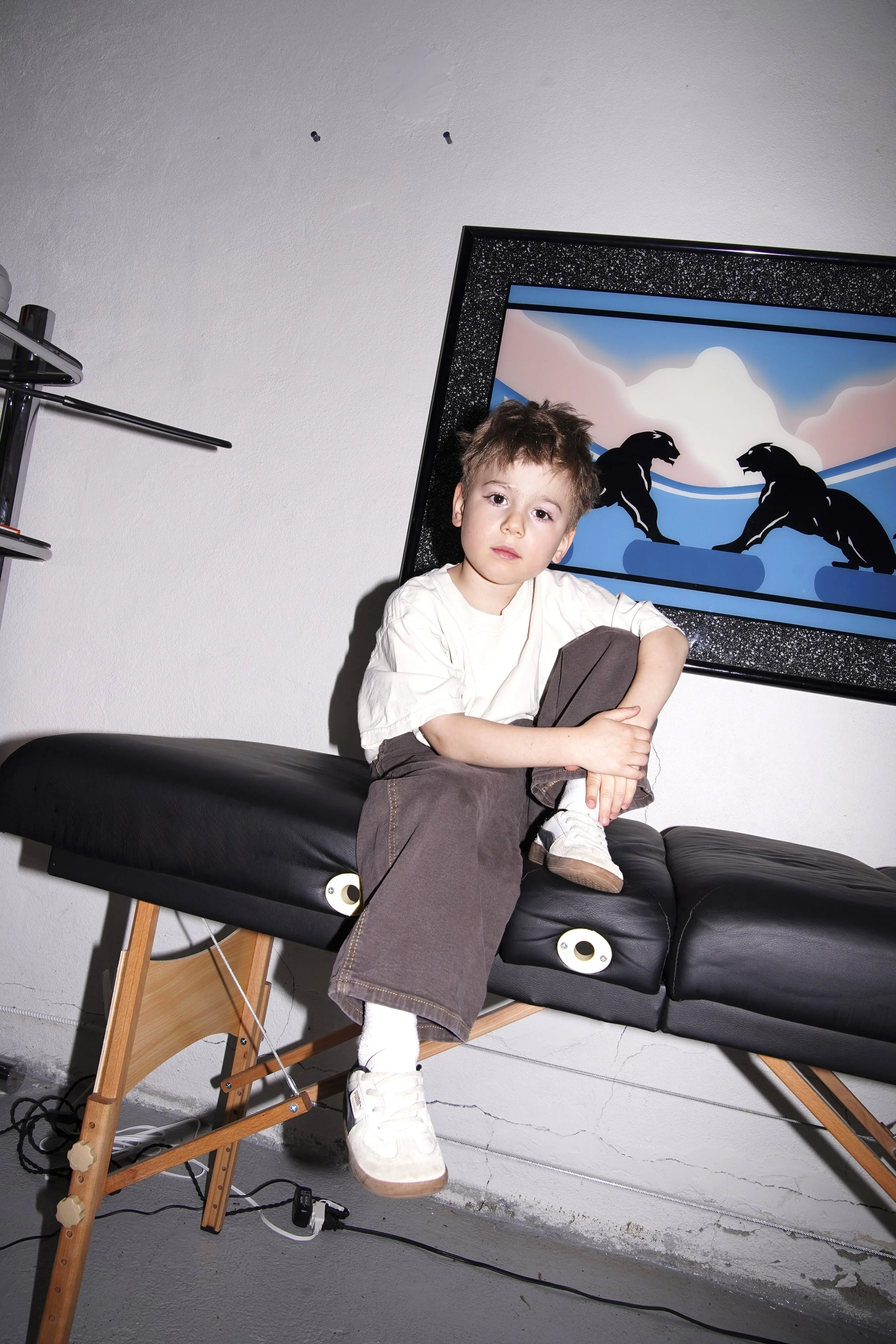 A young boy with tousled hair sitting on a black massage table, wearing a white shirt, brown pants, and white sneakers, with one leg crossed over the other. Behind him, a framed artwork depicts two black cats facing each other against a pink and blue background.