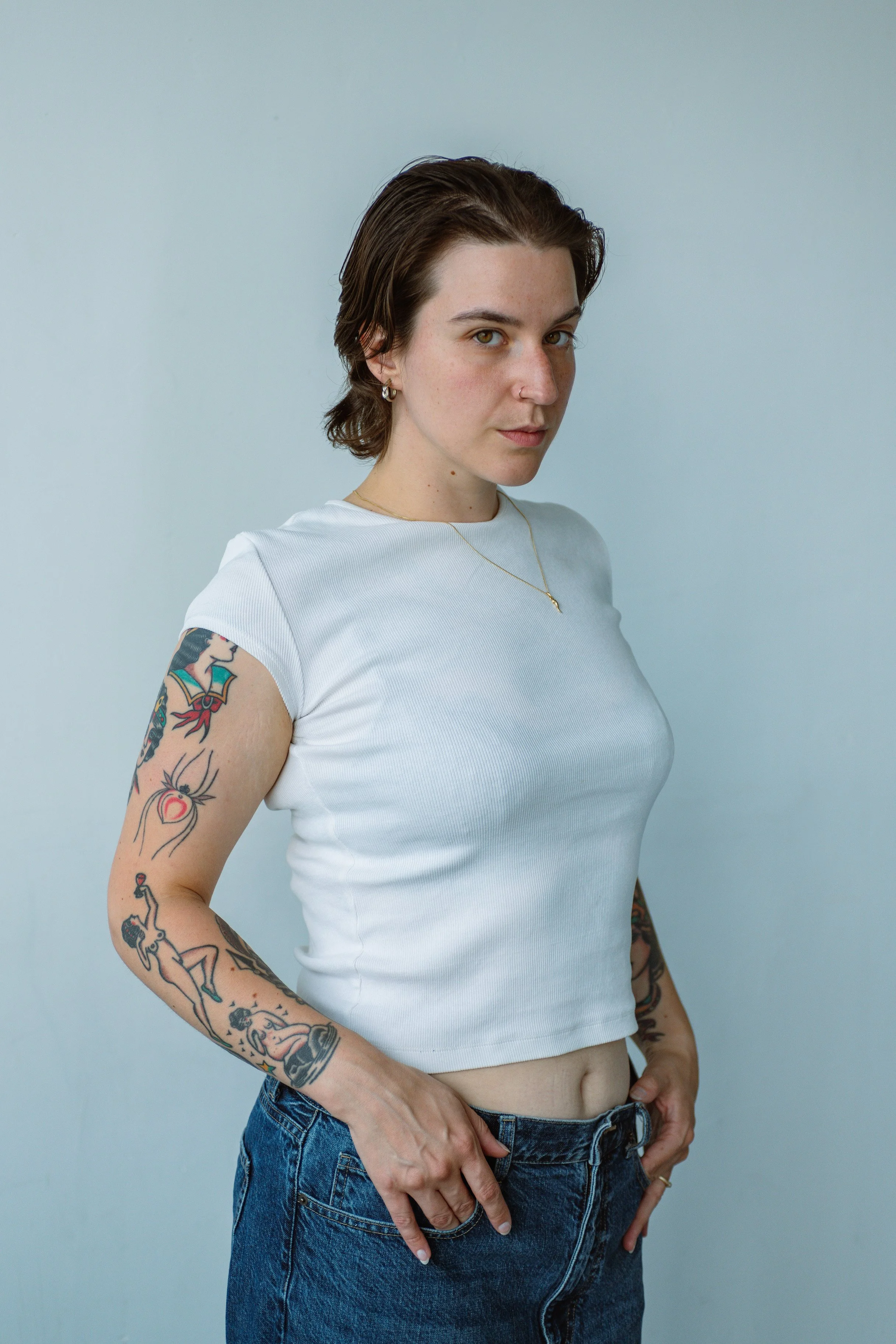 Portrait of a young woman with short brown hair wearing a white crop top and blue jeans, standing against a plain background.