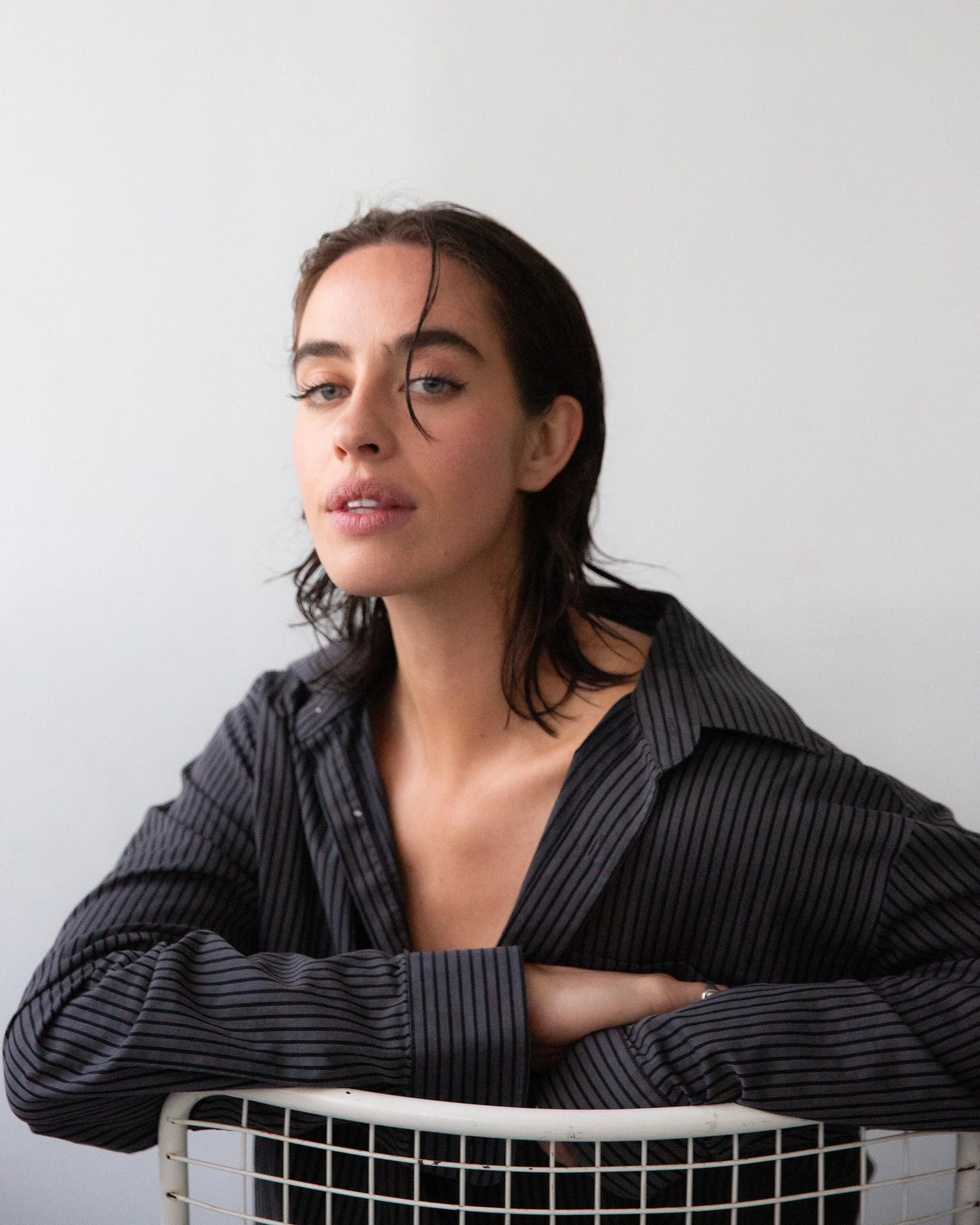 A woman with dark, wet hair and light skin, wearing a black pinstripe shirt, leaning on a white wire chair against a plain light gray wall.