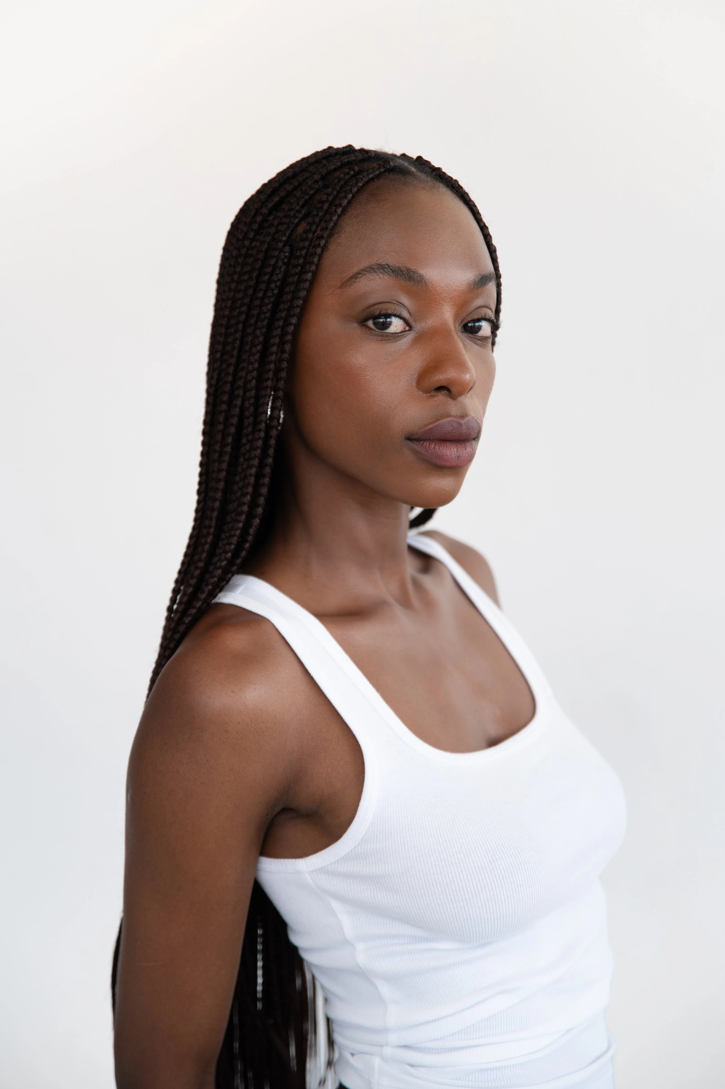 A woman with long braided hair wearing a white tank top, posing against a plain light background.