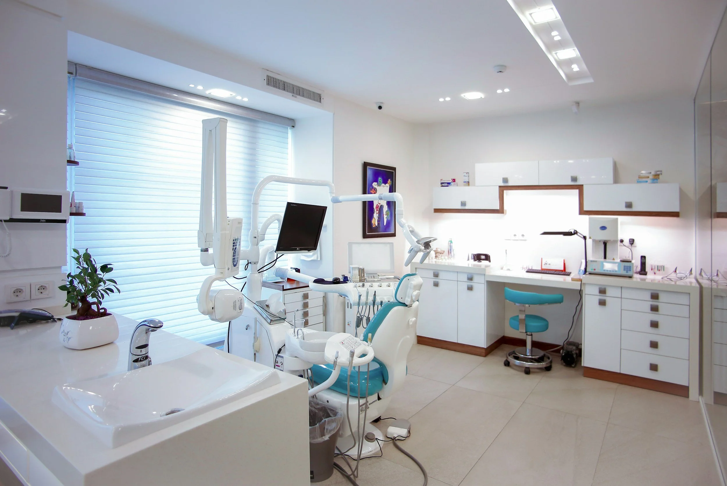 A modern dental clinic room with dental chair, equipment, white cabinets, and a window with blinds.