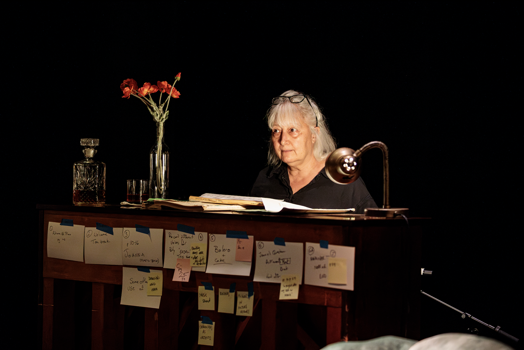 An elderly woman with glasses on her head sitting at a desk, reading a book with notes and pages, in a dark room with a black background. The desk has a bottle and a vase with red flowers, a lamp, and colorful sticky notes attached to the front.