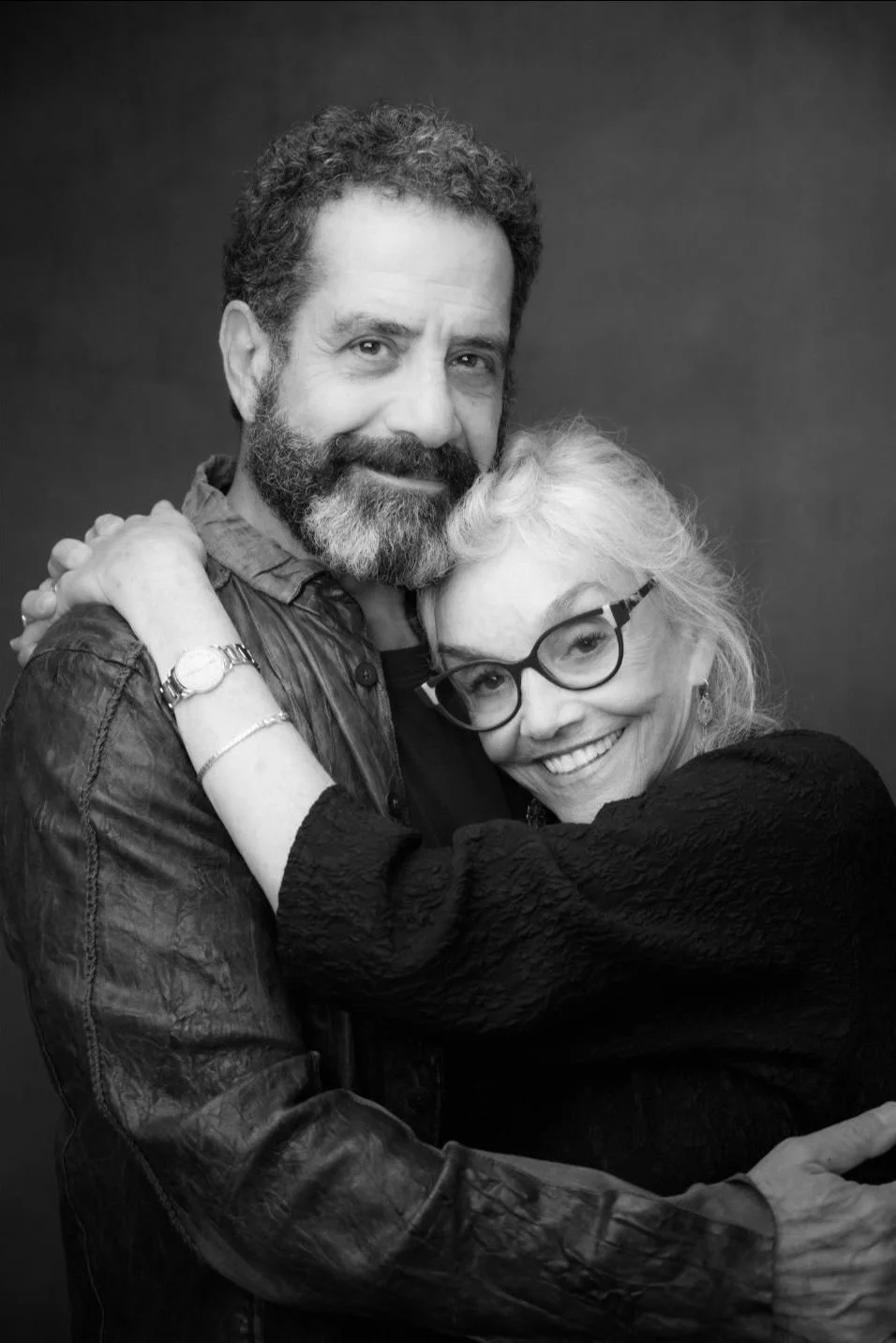 A black and white photograph of a man and a woman embracing and smiling at the camera. The man has curly hair and a beard, and is wearing a leather jacket. The woman has light-colored hair, glasses, and is wearing a textured black top. They appear to be happy and close.