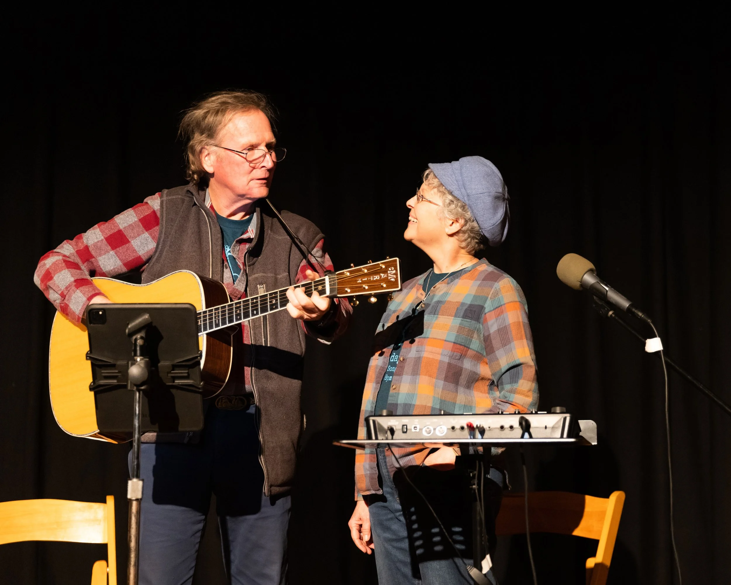 A man playing guitar and an elderly woman singing into a microphone on a black stage with wooden chairs and music equipment.