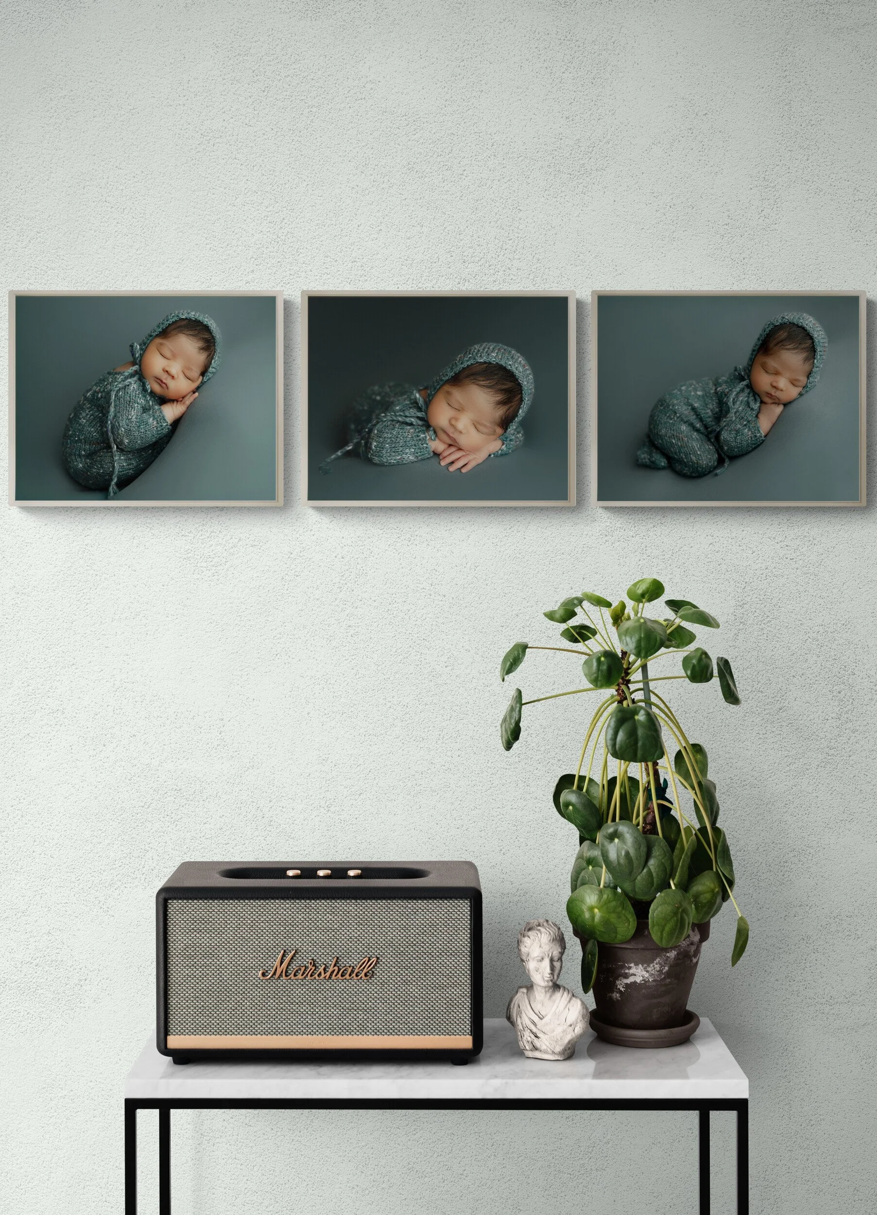 A tabletop with a Marshall speaker, a small bust sculpture, and a potted plant. Above, three framed photographs of a sleeping baby dressed in a knitted outfit are hung on the wall.