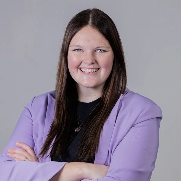 A young woman with brown hair, blue eyes, and fair skin is smiling at the camera. She is wearing a light blue blazer over a cream top, with a small gold necklace, and is standing against a plain light gray background.