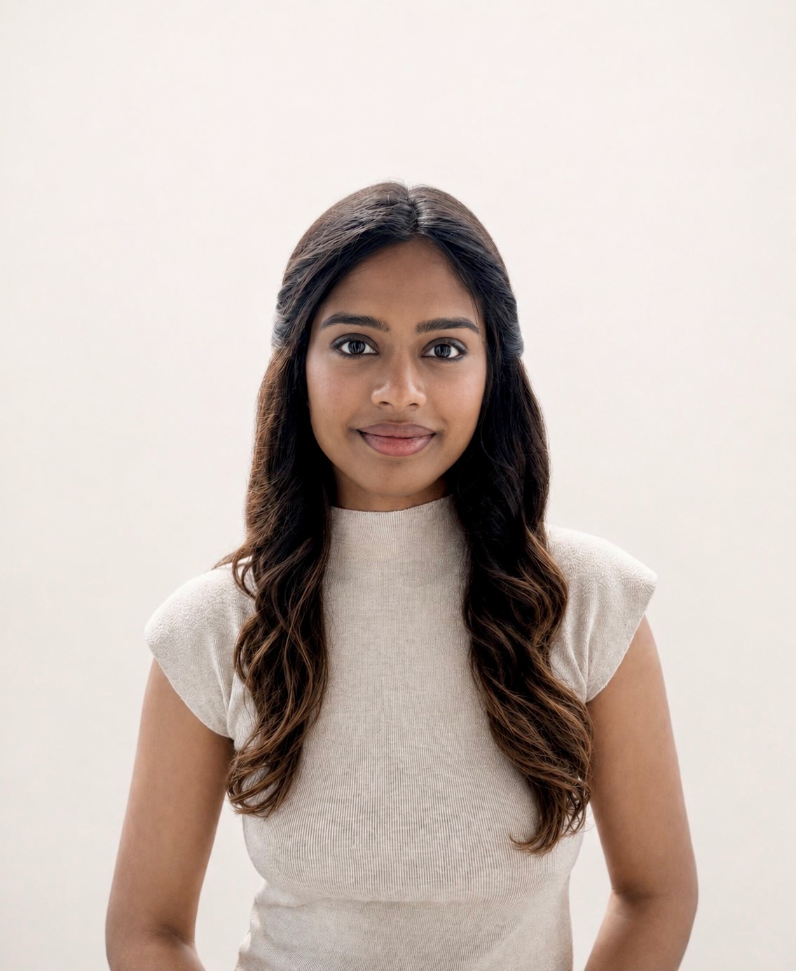 A young woman with long wavy brown hair, wearing a beige sleeveless top, standing against a plain light-colored background.