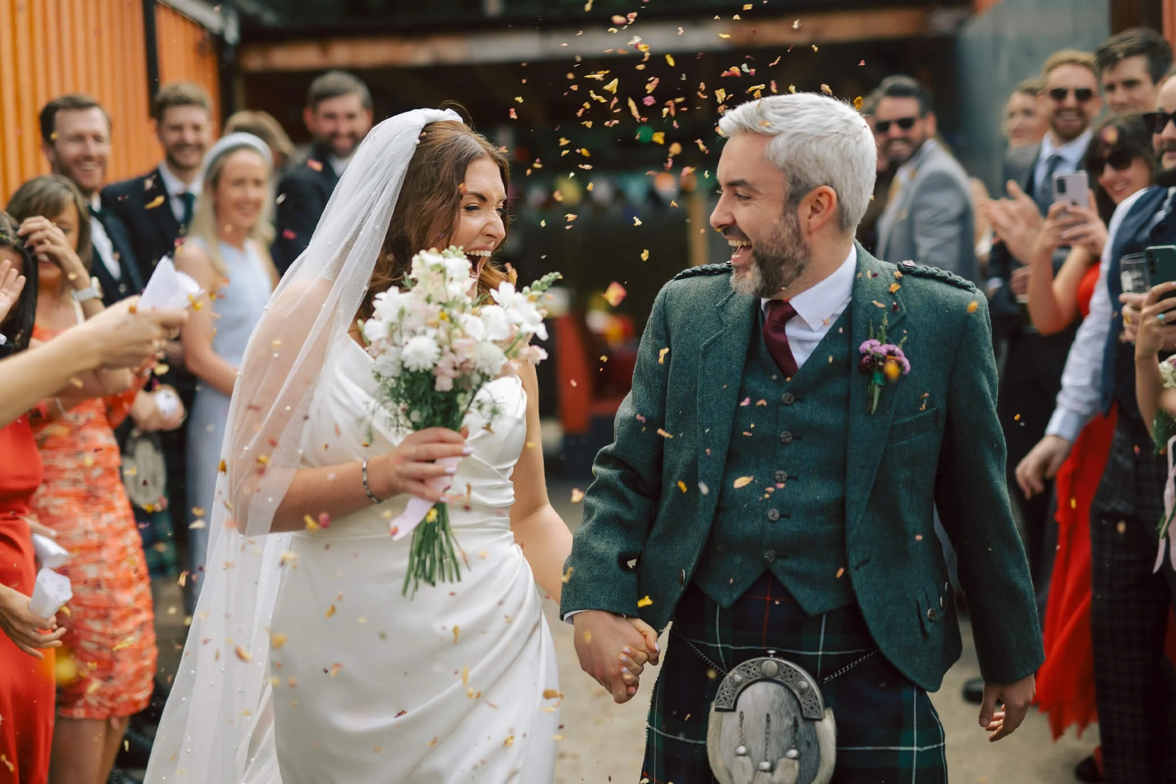 A joyful bride and groom holding hands at their wedding, surrounded by friends and family celebrating with confetti.