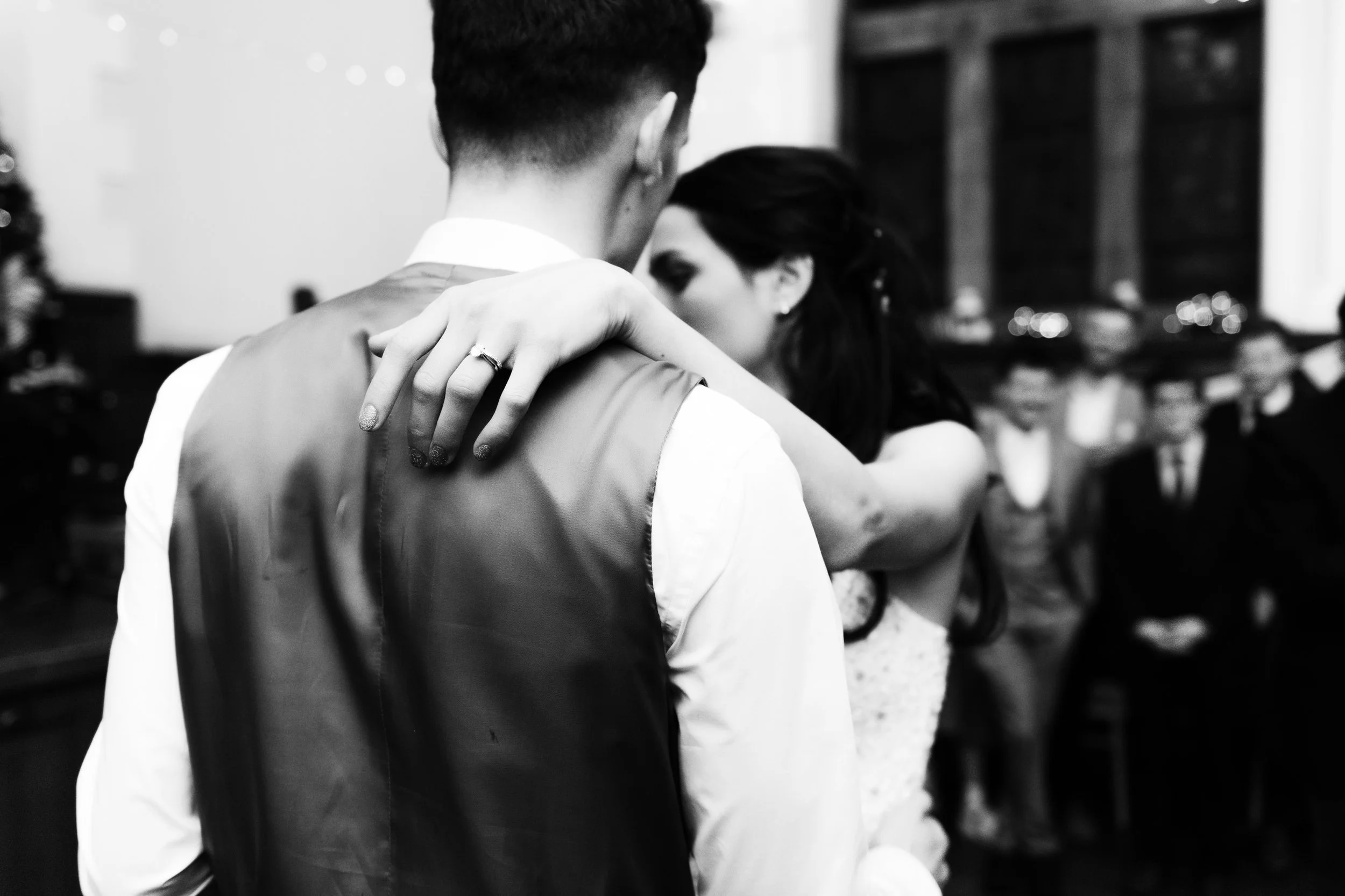 A black and white photo of a couple dancing closely, with the woman's arm around the man's shoulder, at a formal event with other seated guests in the background.