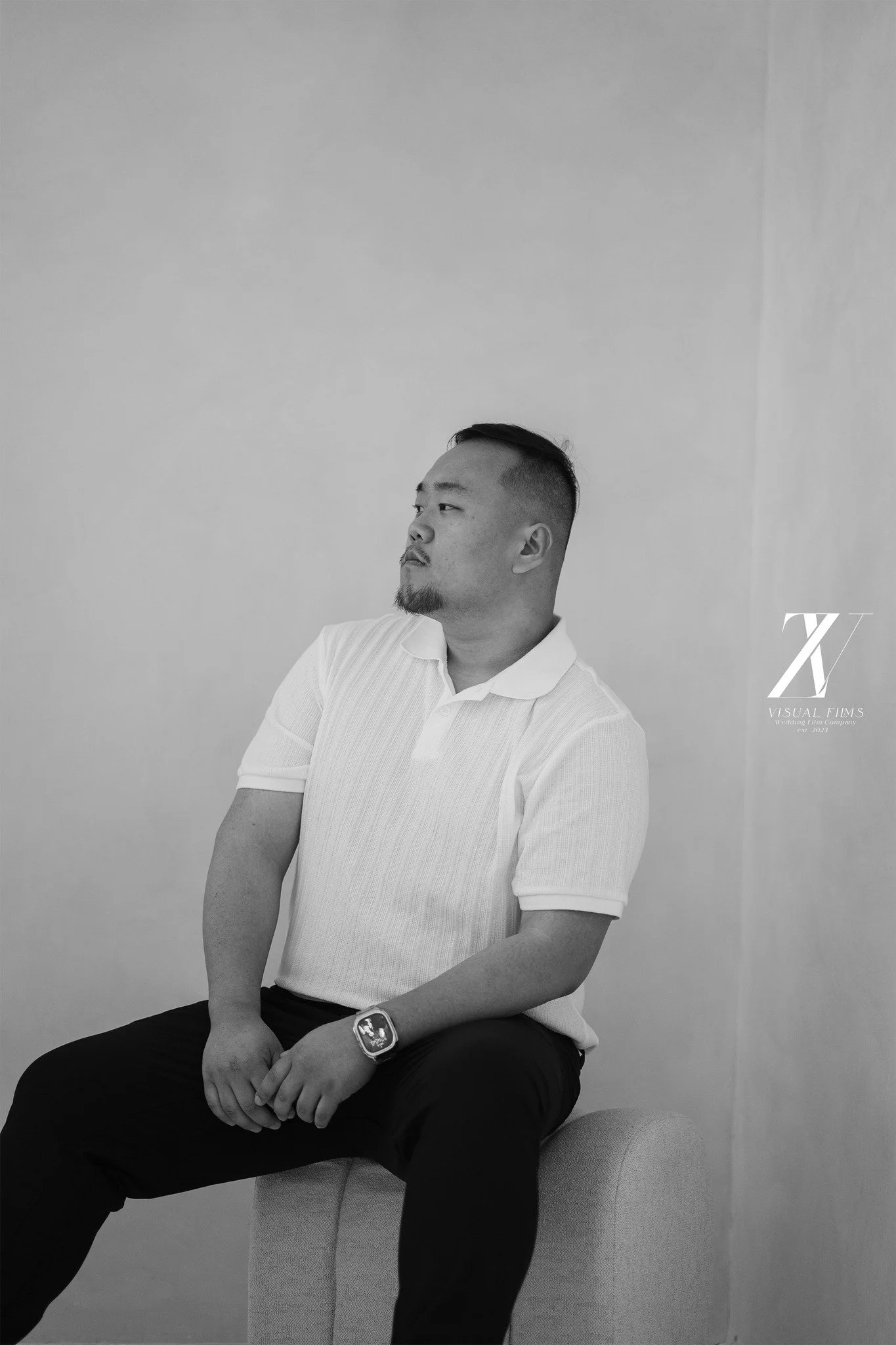 A man sitting on a beige chair against a plain wall, dressed in a white polo shirt and black pants, wearing a smartwatch, with arms resting on his lap, looking to the side.