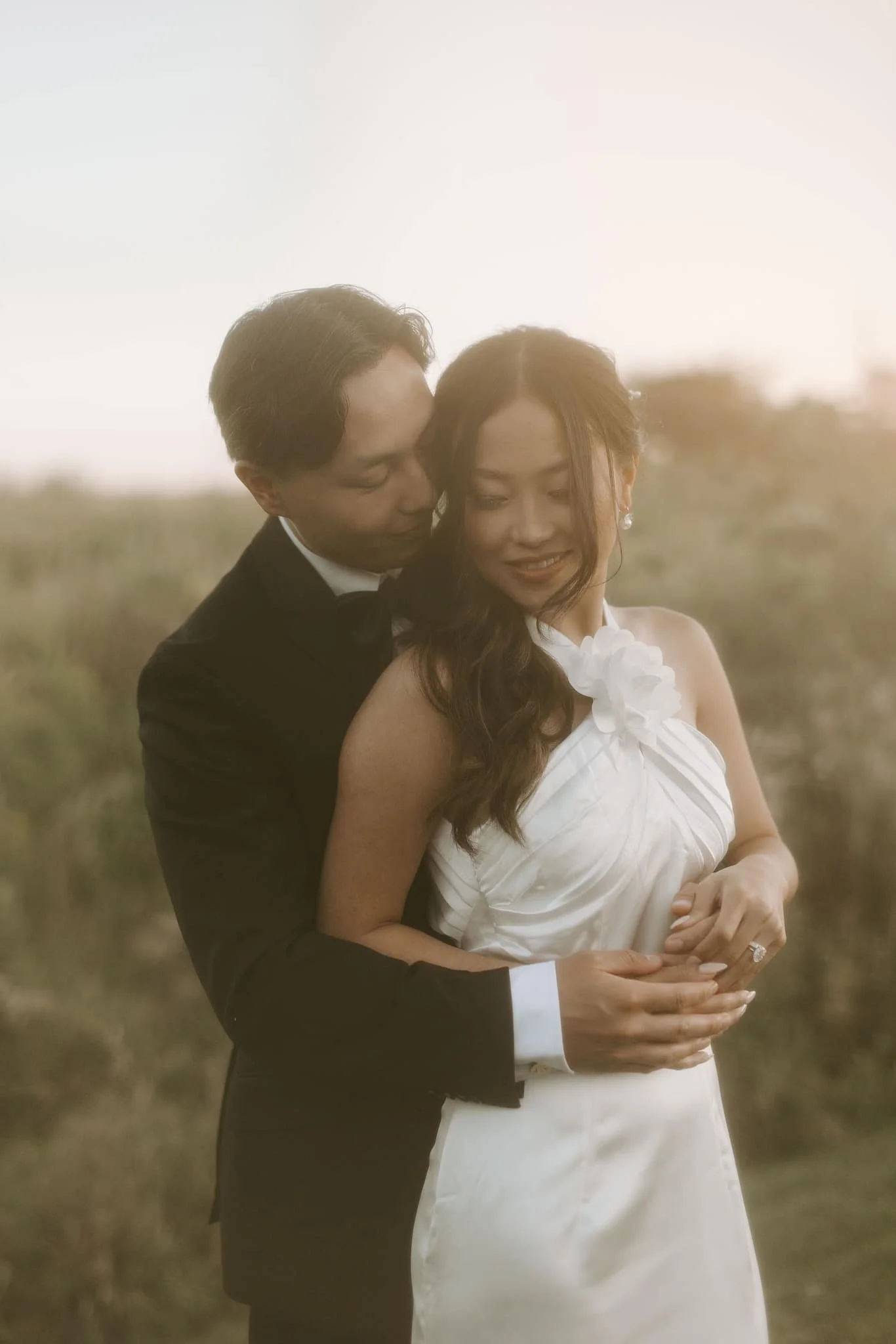 A couple is embracing outdoors at sunset. The man is wearing a black suit and the woman is in a white dress. They are smiling and close together.