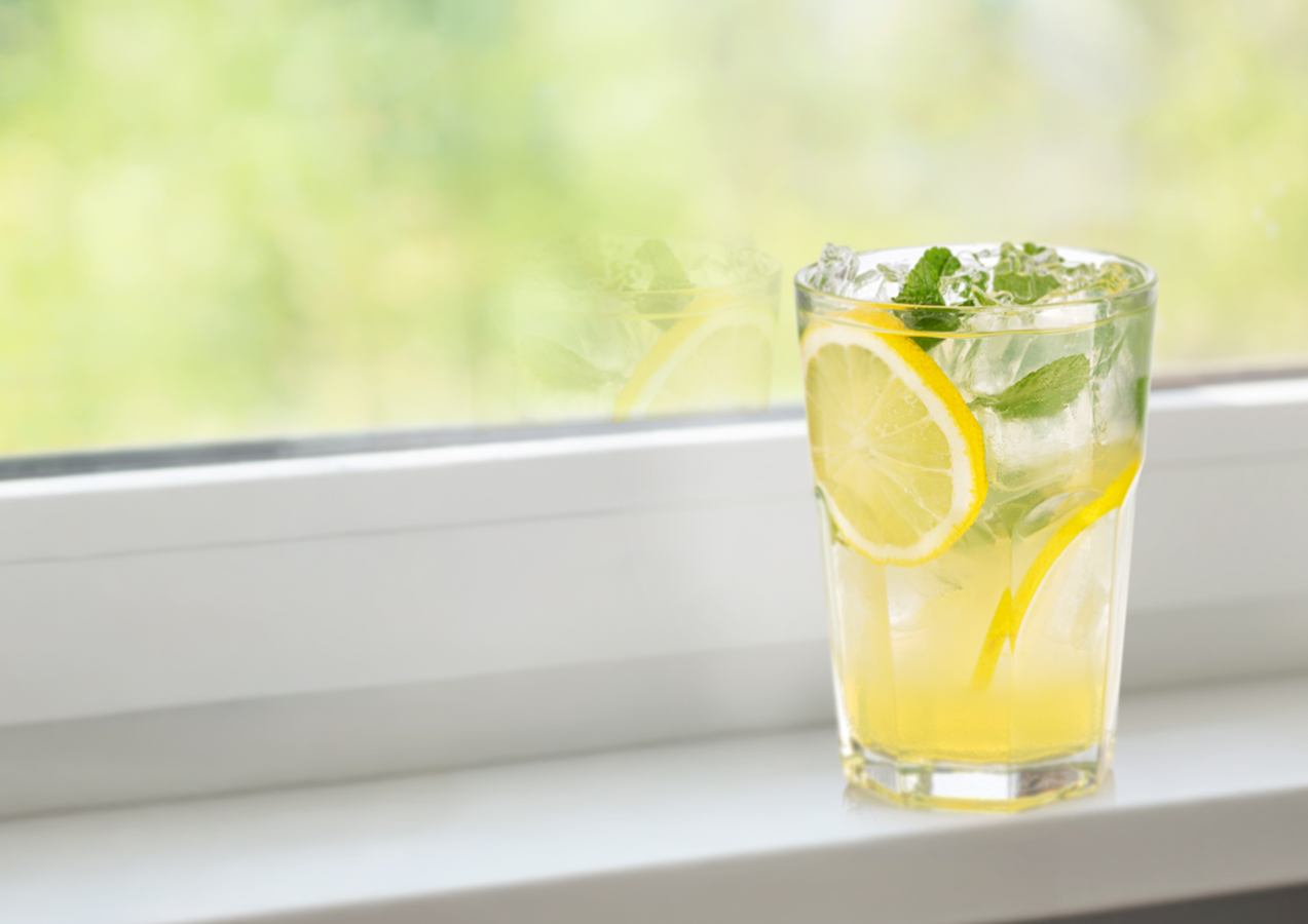 A glass of lemon and mint beverage with ice on a windowsill.