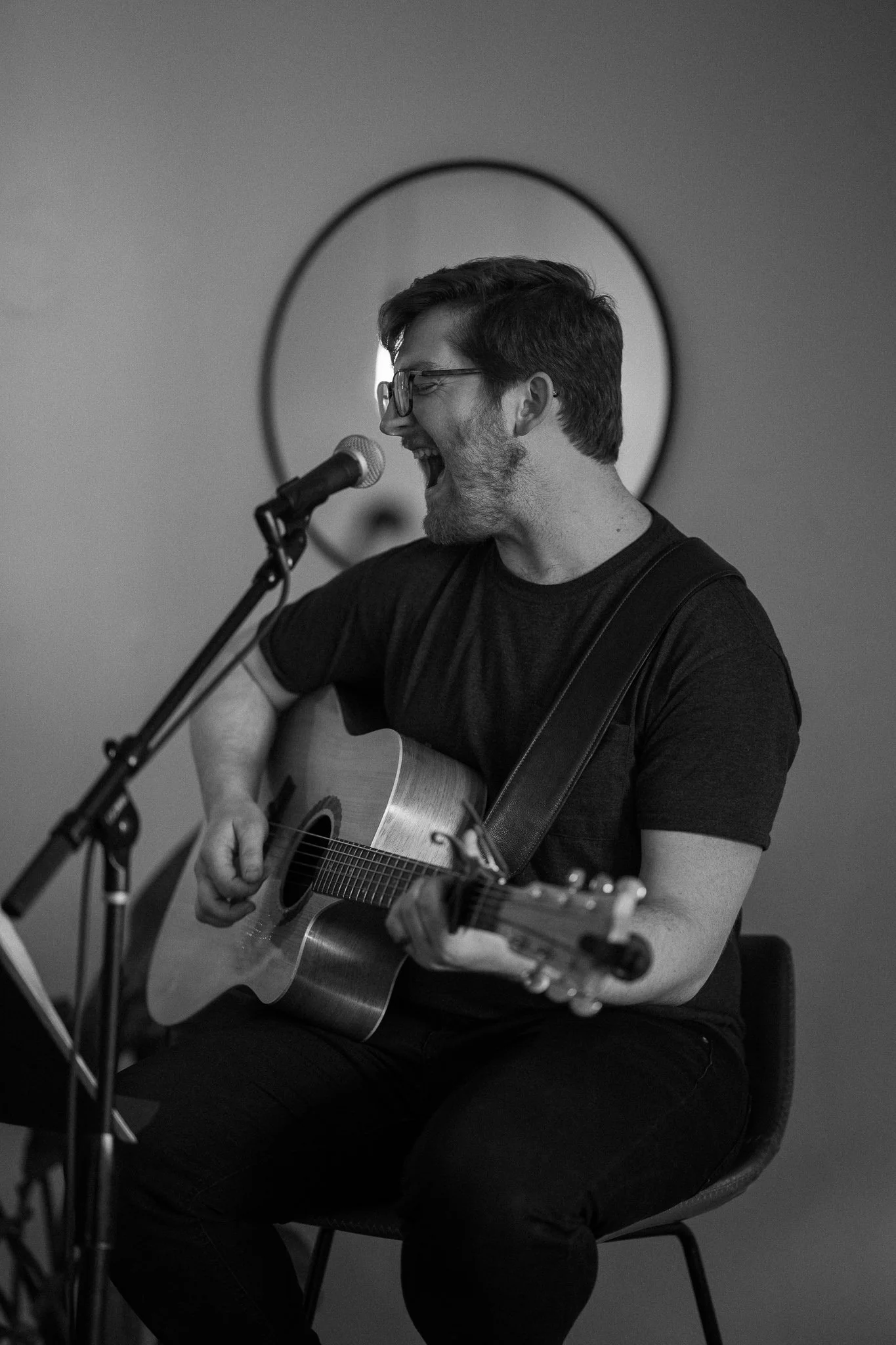 Black and white photo of a person playing an acoustic guitar and singing into a microphone, sitting on a chair with a round wall mirror in the background.