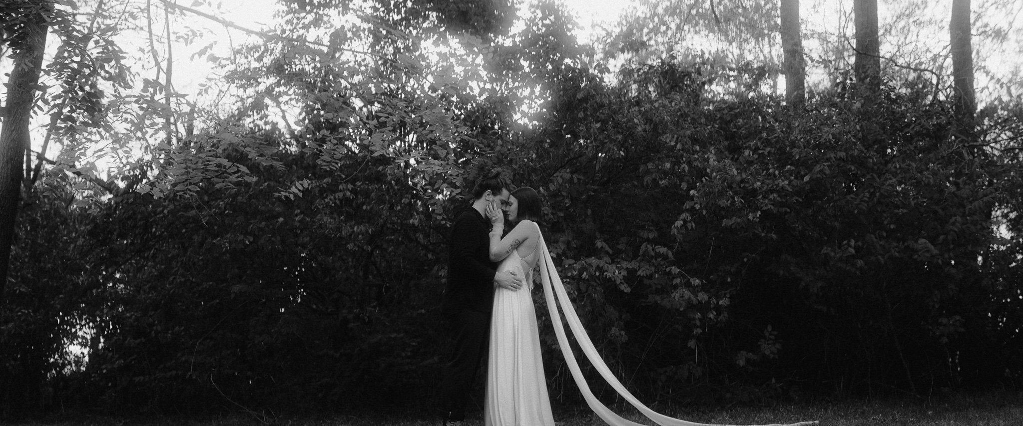 Romantic couple embracing in a forest setting, bride in flowing gown, black and white photo.