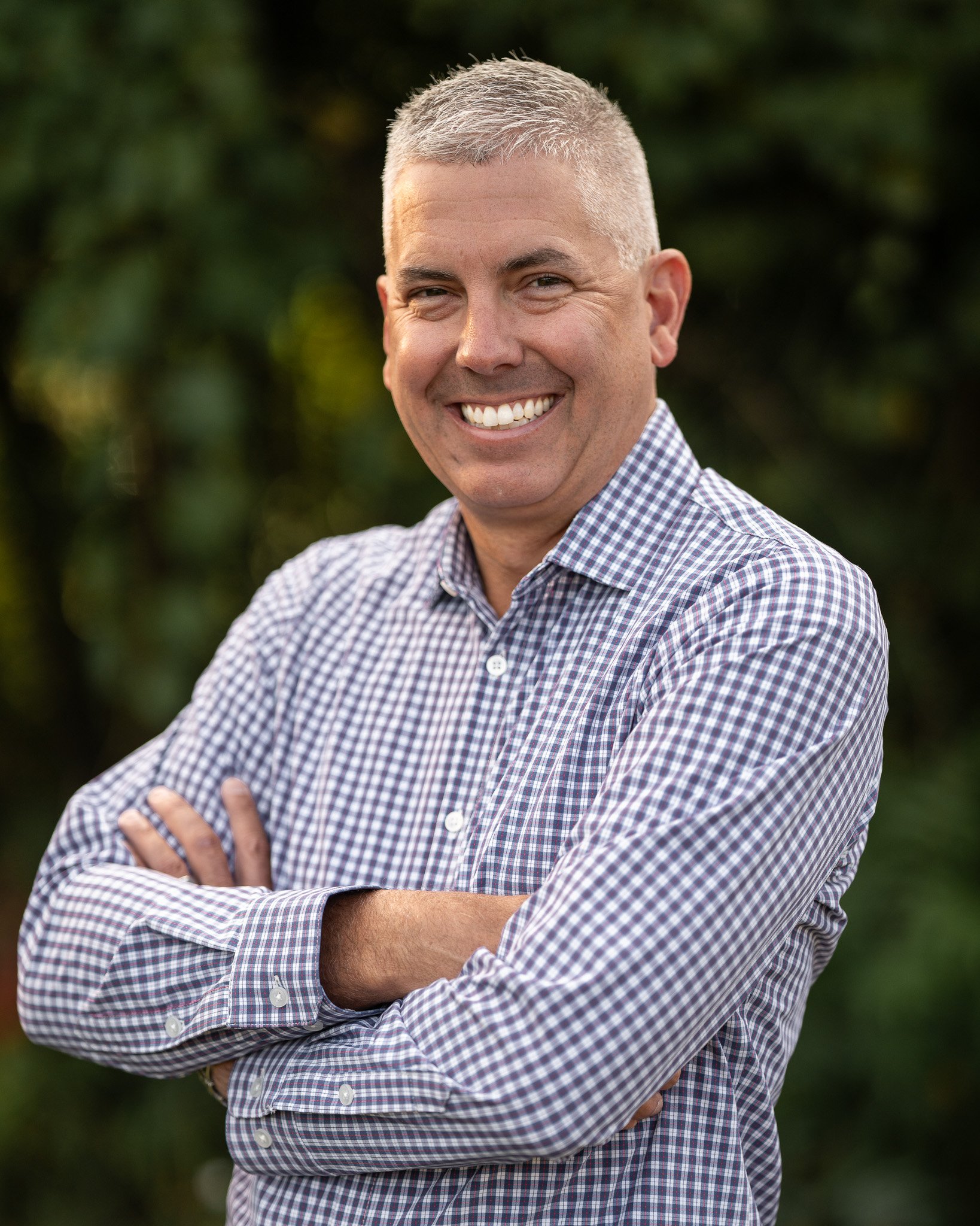 Smiling man with short gray hair wearing a checkered shirt, arms crossed, standing outdoors.