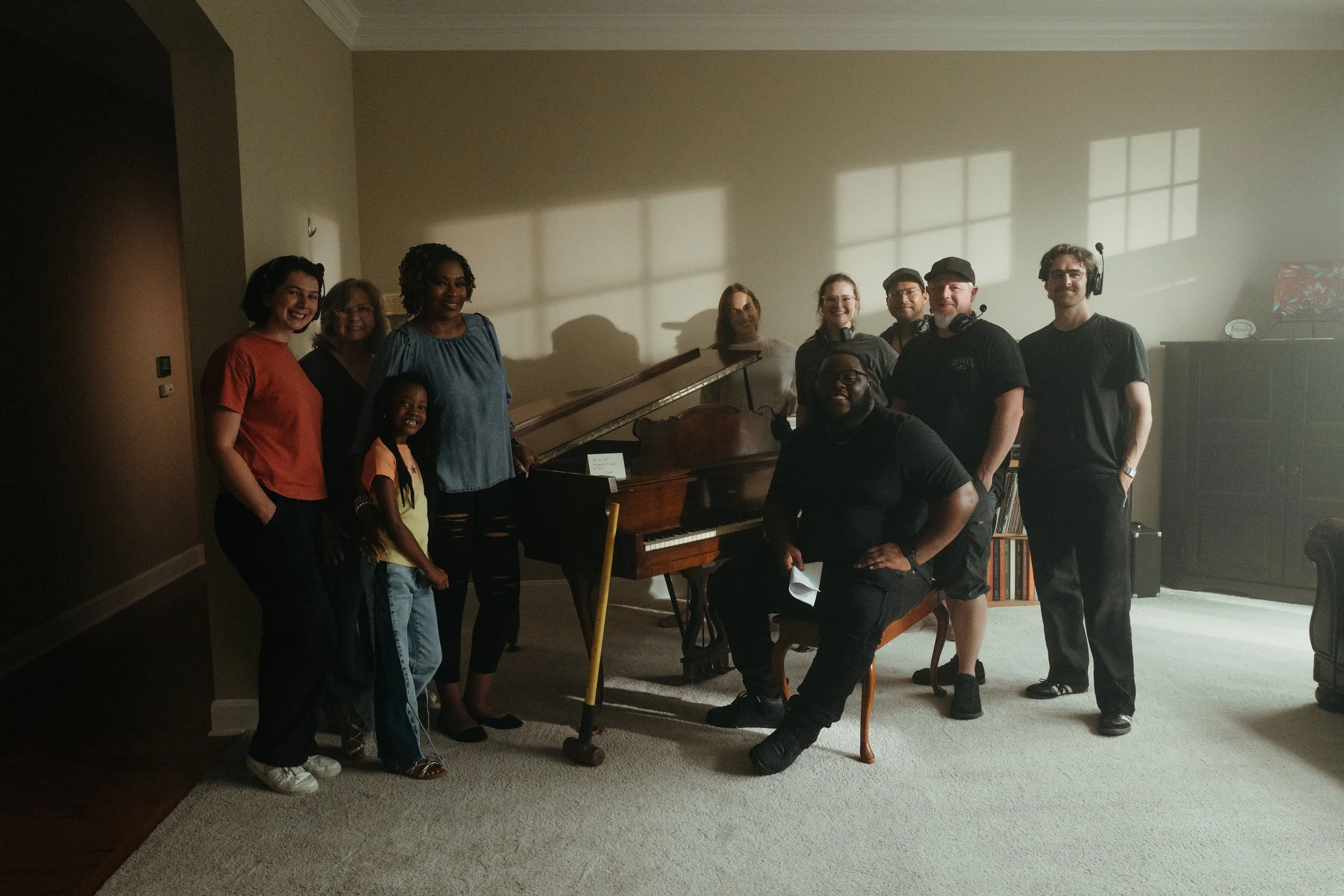 A group of ten people, including children and adults, posing together in a living room near a piano. The sunlight creates grid-like shadows on the wall behind them, and a music stand with a floral-patterned cover is visible on the right side.