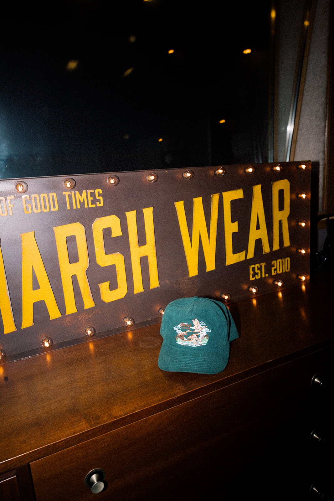A wooden sign with illuminated lights displaying the words "MARSH WEAR EST. 2010" in yellow, placed on a dark surface. A teal cap with a fish embroidery is in front of the sign.