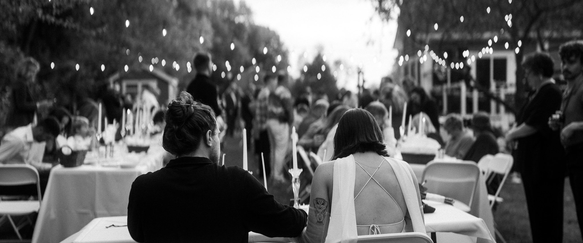 Black and white photo of an outdoor gathering with people seated at tables adorned with candles and string lights overhead.