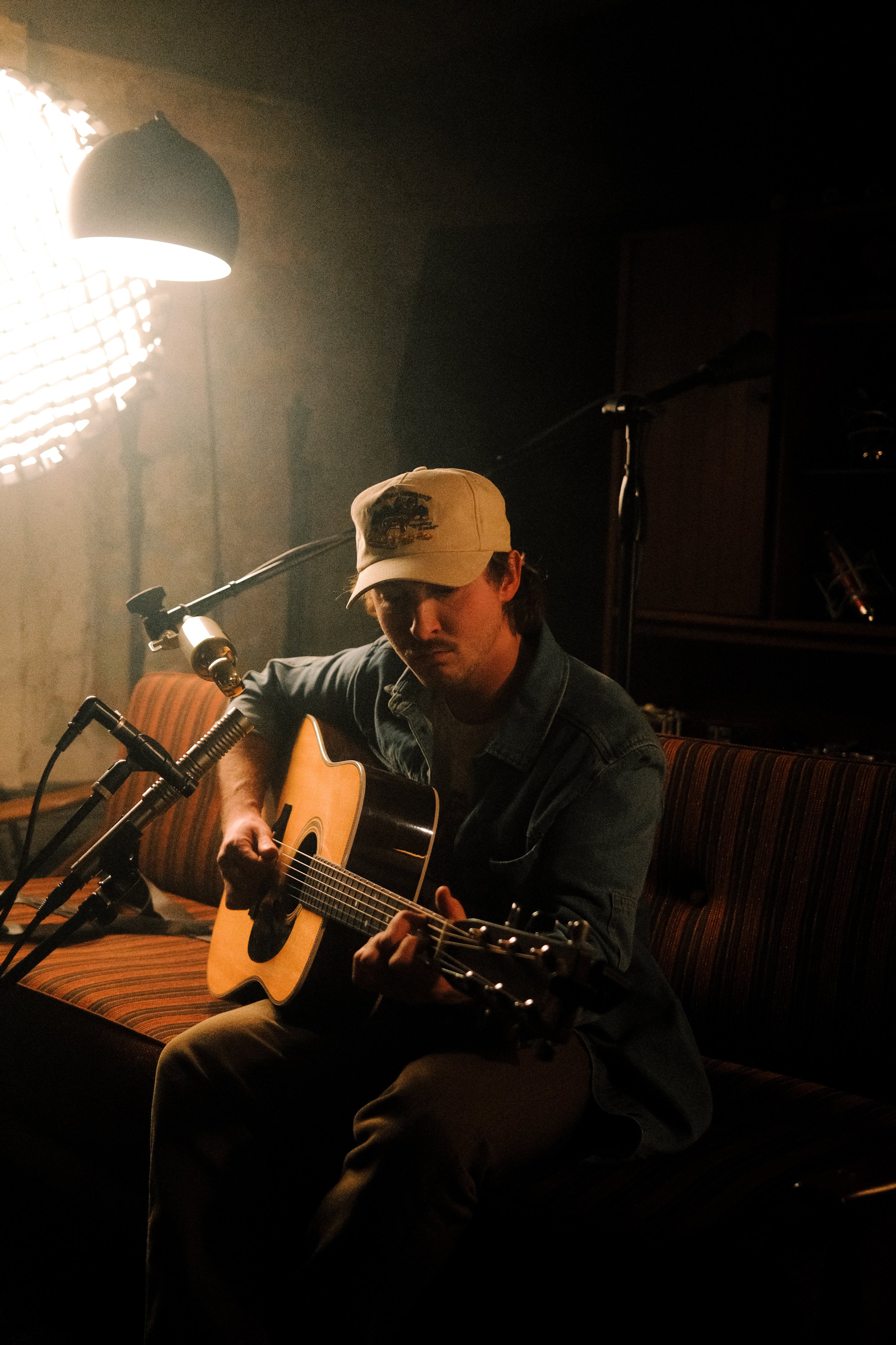 Person playing an acoustic guitar in a dimly lit room with a microphone and soft lighting.