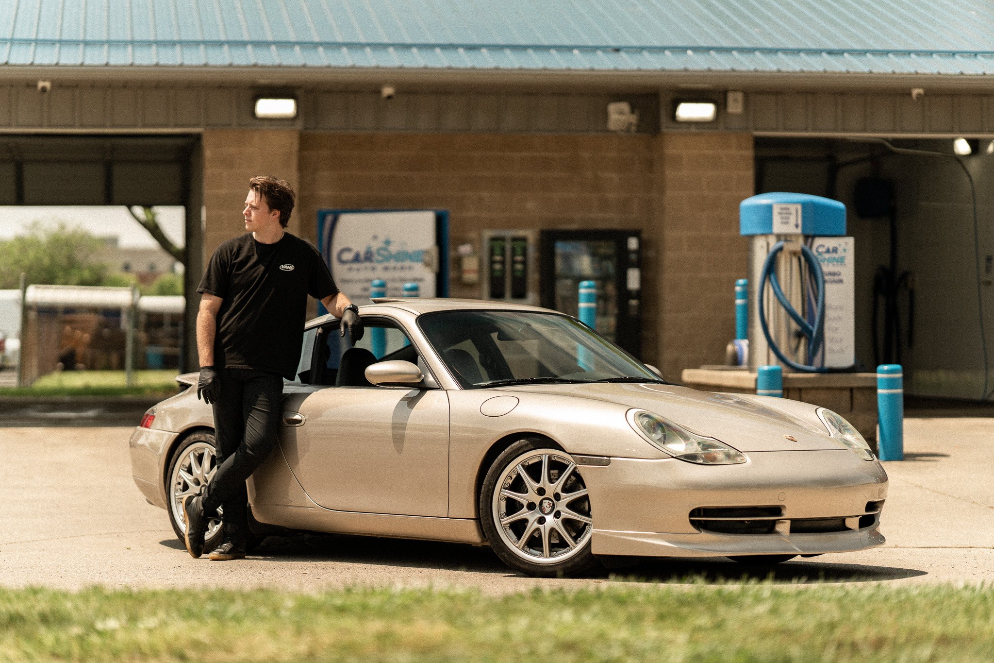 Person standing next to a gold Porsche at a car wash, wearing a black shirt and gloves.