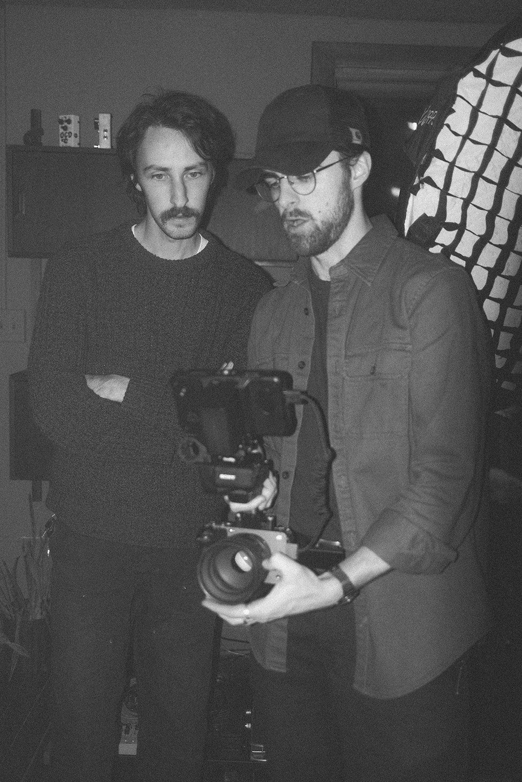 Two men examining a camera setup; one holds the camera, and the other observes, both in a dimly lit room.