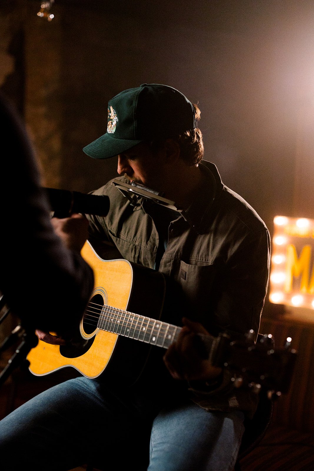 Musician wearing cap playing guitar with harmonica on stage, dimly lit atmosphere.