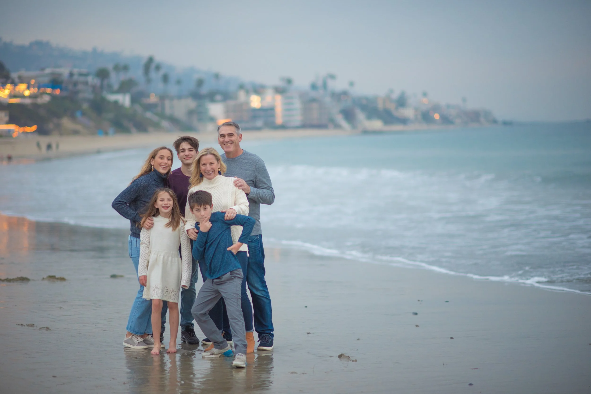 Family of seven standing on the beach near the water at dusk, smiling and posing for a photo. Beach and residential buildings are visible in the background.