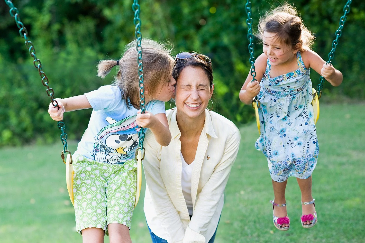 A woman playing on a swing with two young girls, all smiling and enjoying outdoors in a park with green grass and trees in the background.