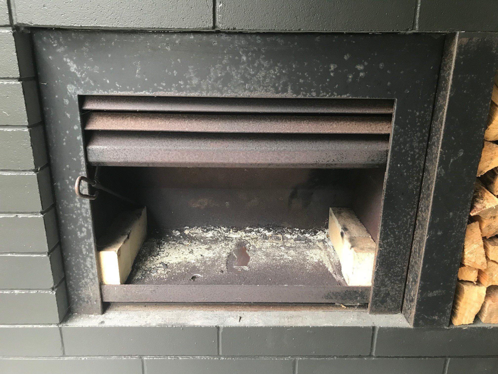 Close-up of a black outdoor fireplace or grill with a metal vent, with firewood stored to the side and stone below.
