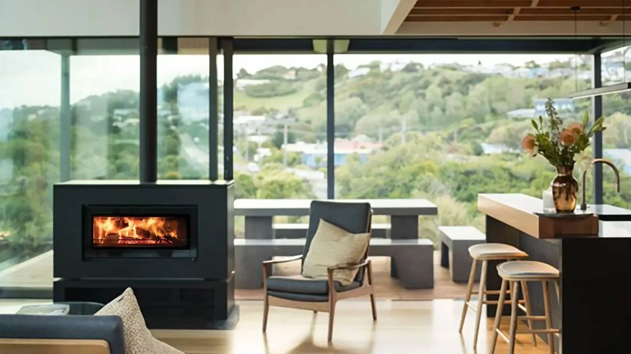 Interior of a modern living room with large floor-to-ceiling windows showing a hilly landscape outside; a lit fireplace on the left, a wooden armchair with a cushion in front of the fireplace, a counter with a flower vase on the right, and bar stools.