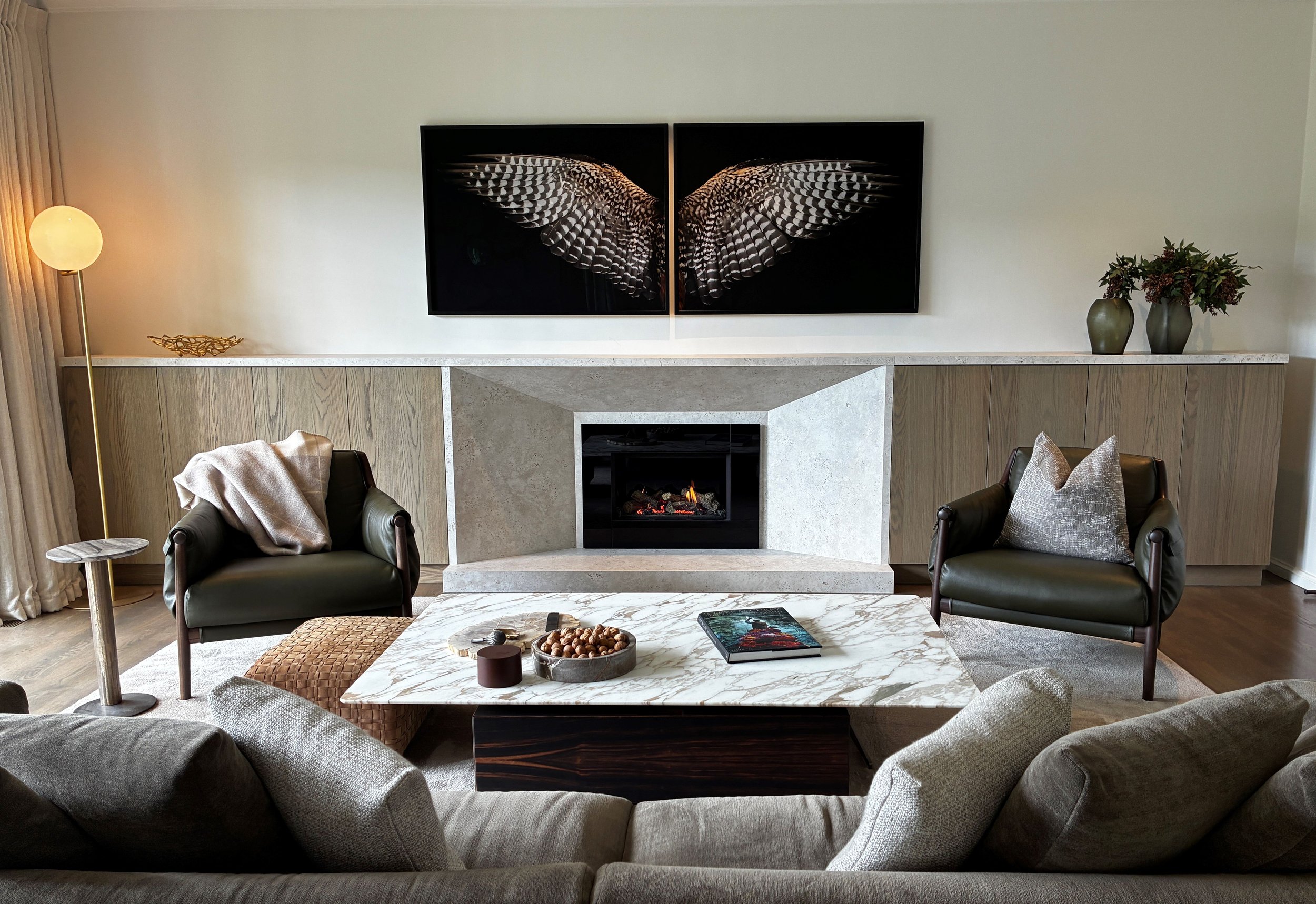 Living room with stone fireplace, two black armchairs, a marble coffee table, a side table with a blanket, a floor lamp, a wall-mounted art piece of butterfly wings, and vases with flowers.
