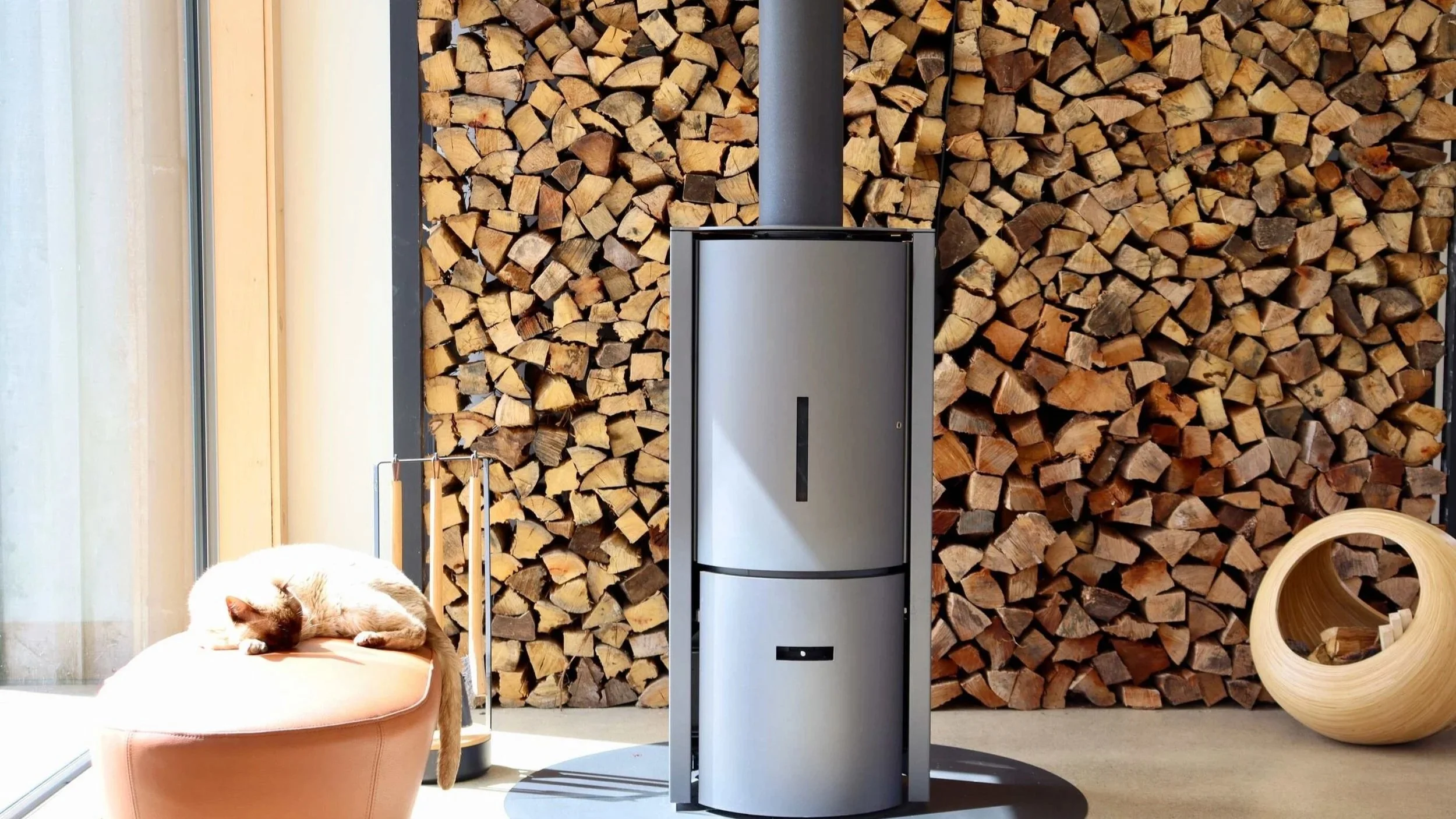 A cat sleeping on an orange ottoman in front of a wall of stacked firewood, with a modern wood-burning stove in the center and a wooden circular sculpture on the right.