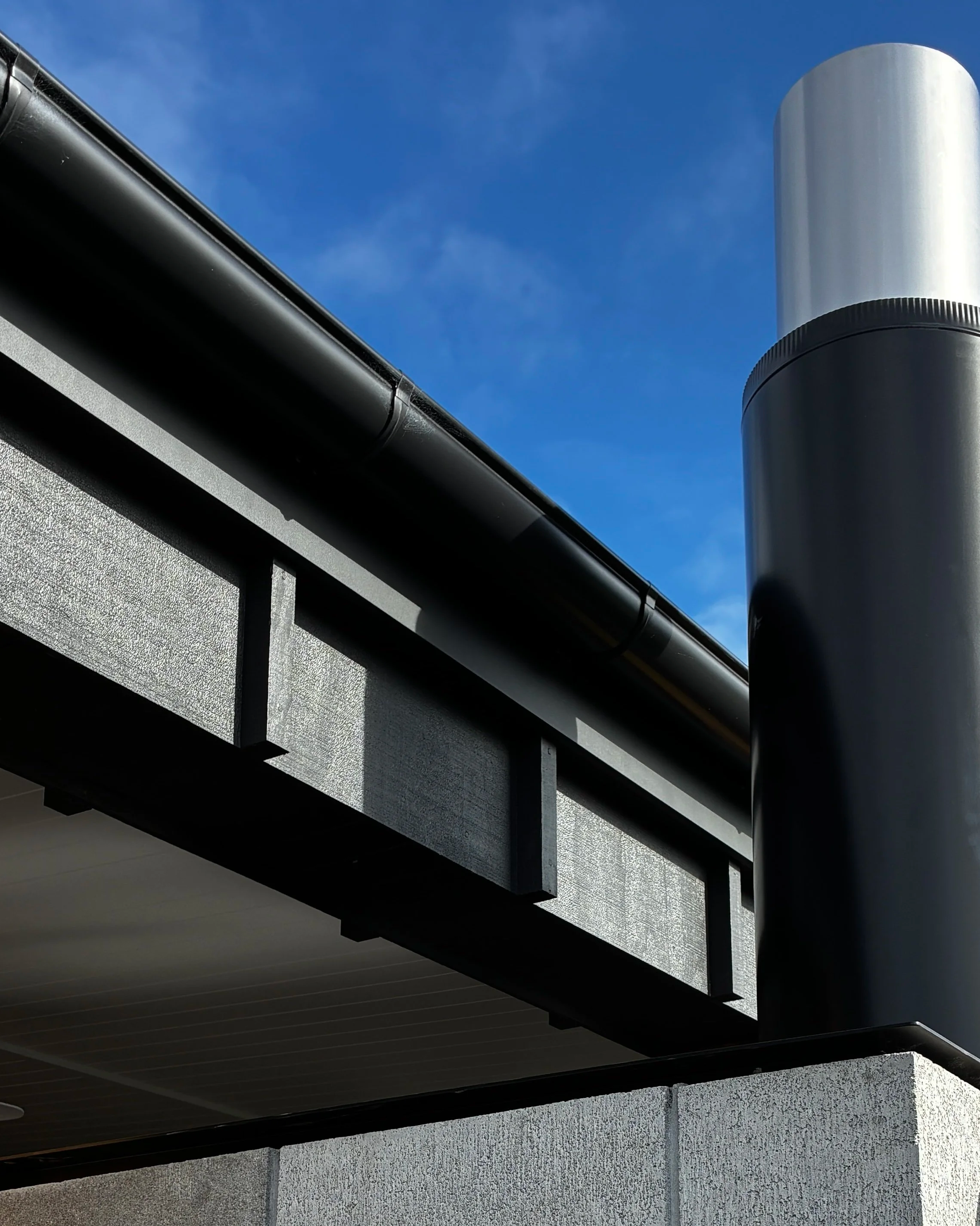Close-up of a modern building's exterior with black gutter and downspout, gray textured wall, and sections of black metal with wood paneling beneath, against a bright blue sky.